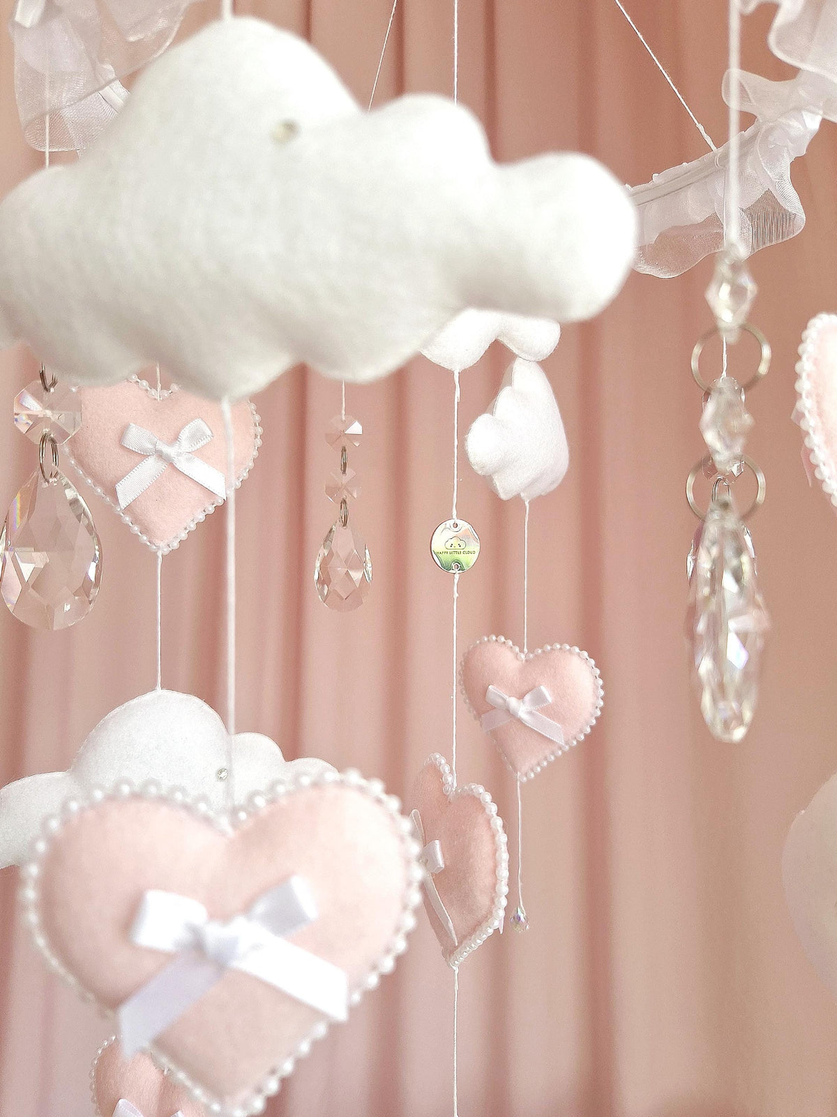 A hanging mobile featuring white felt clouds and pale pink felt hearts suspended by white strings. The hearts are decorated with small white bows and pearl-like beaded edges. Clear, faceted crystal-like teardrop pendants hang between.