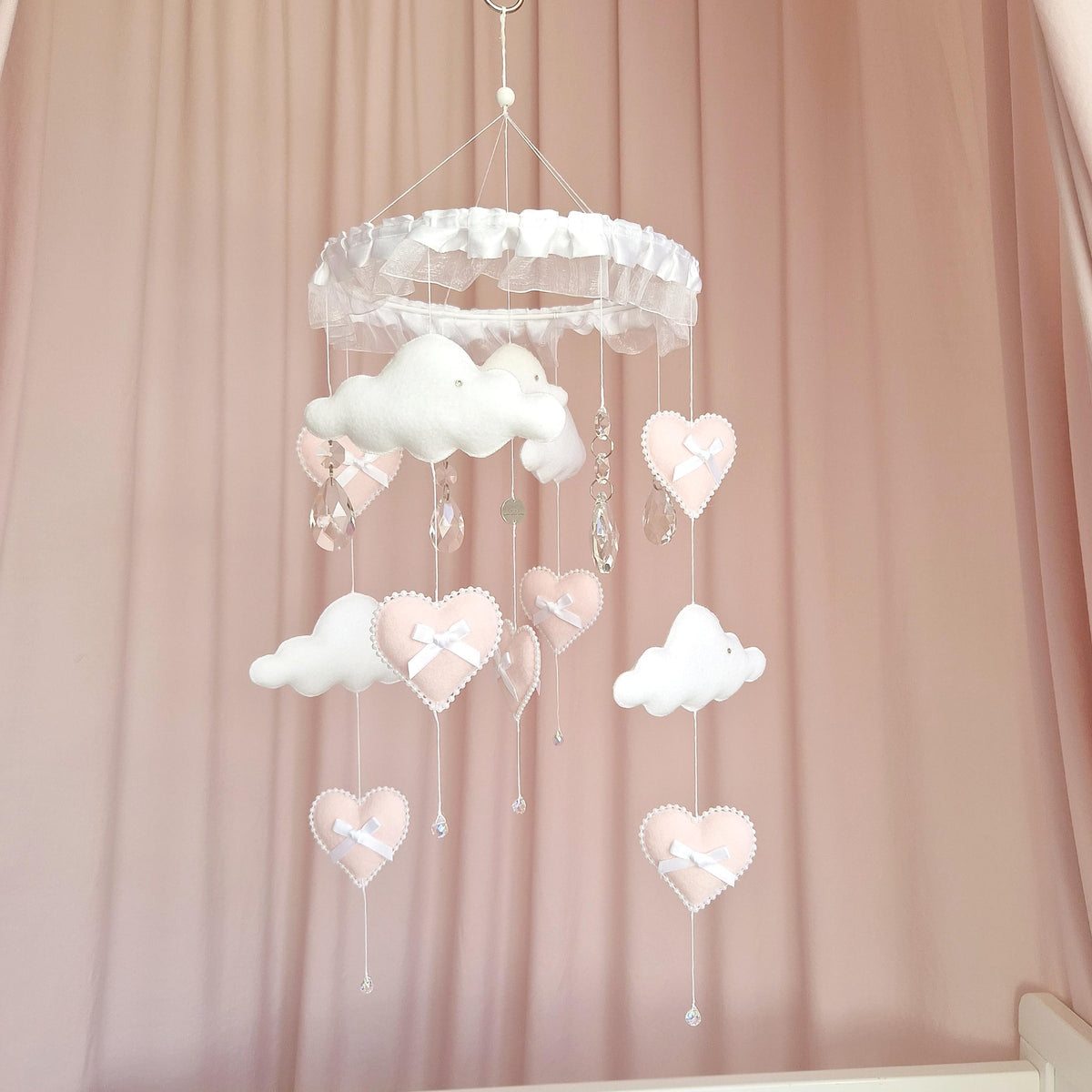 A hanging mobile featuring a white ruffled circular frame with dangling white felt clouds and soft pink felt hearts. Each heart is accented with a small white ribbon bow and white beaded trim. Clear crystal-like pendants hang throughout.