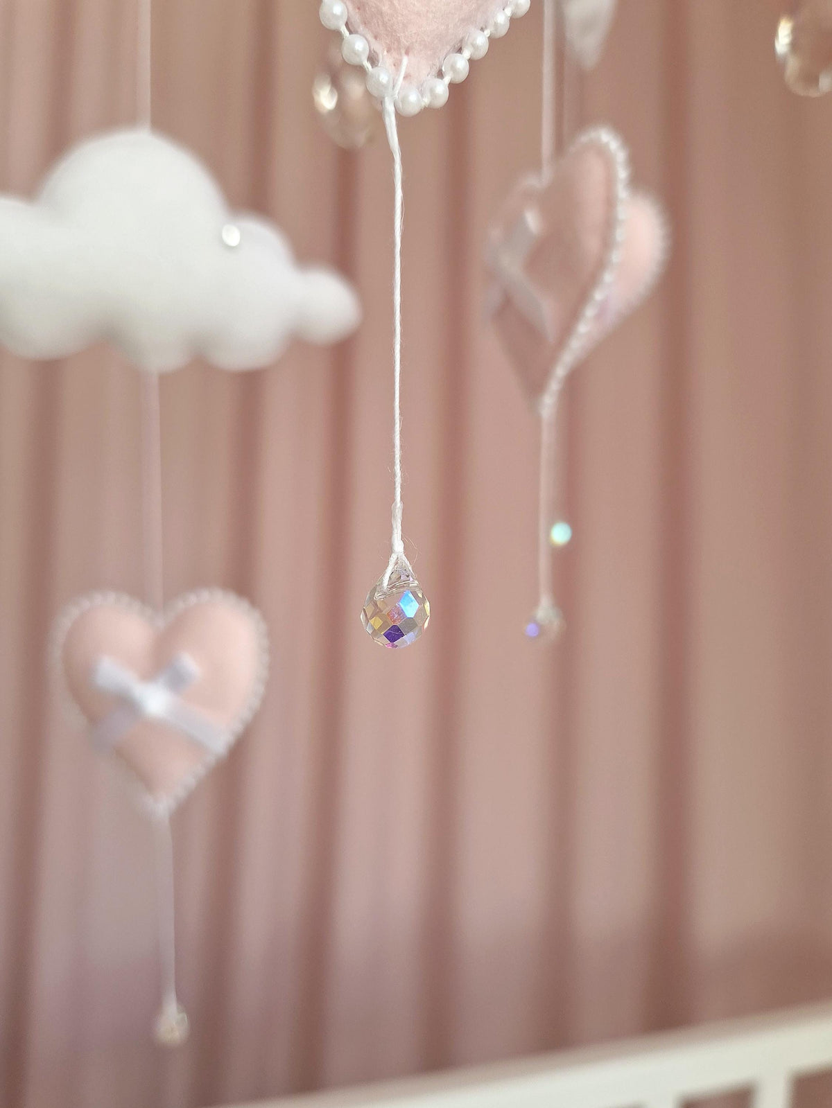 A hanging mobile featuring soft pink felt hearts and white felt clouds suspended by white strings. The hearts are decorated with white ribbon bows and pearl-like bead trim. Clear, faceted crystal-like beads hang from the centre.