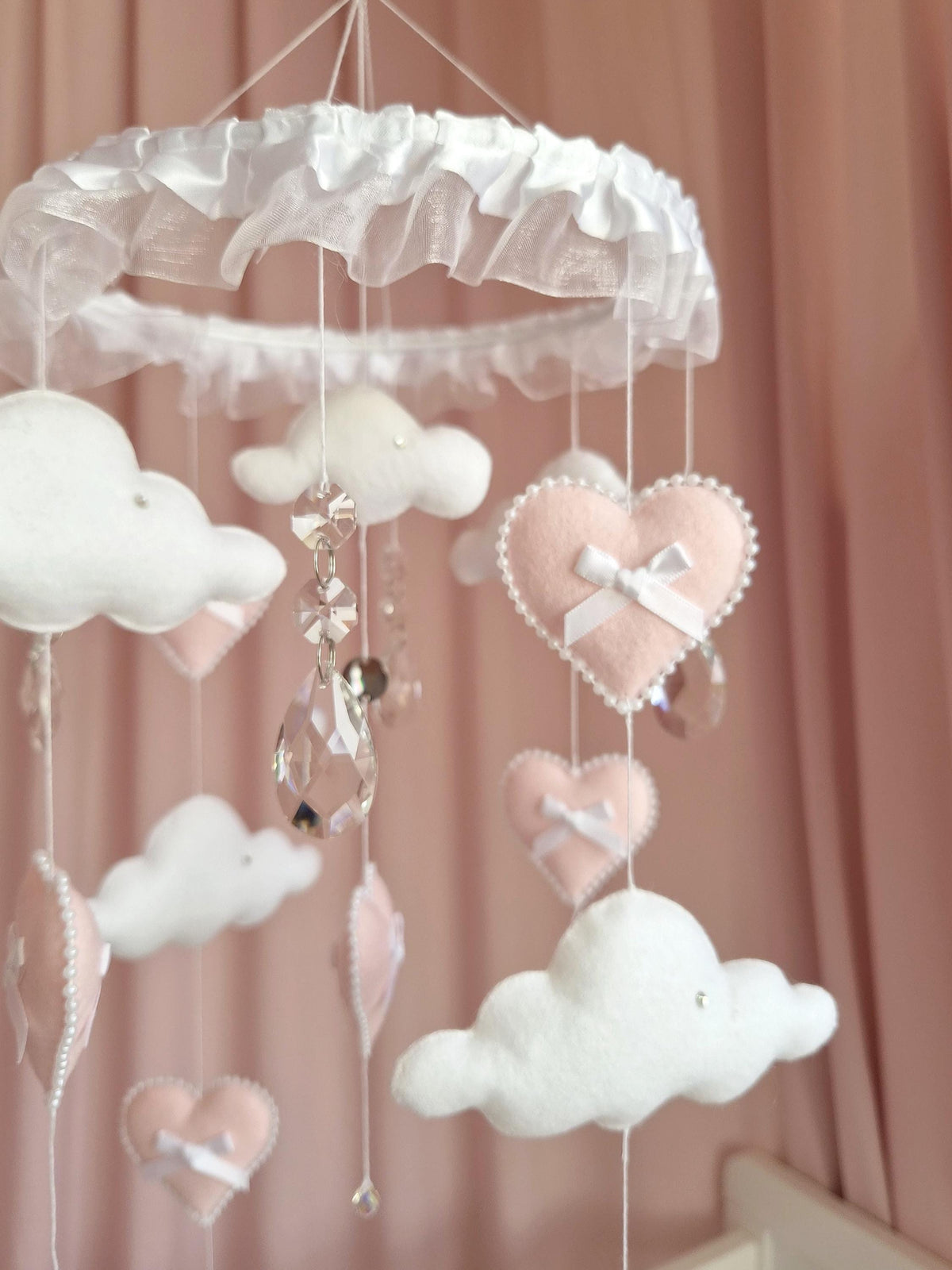 A hanging mobile featuring white felt clouds and soft pink felt hearts suspended from a circular frame with white ruffled fabric. The mobile includes clear faceted crystal teardrop accents and small white bows on each heart.