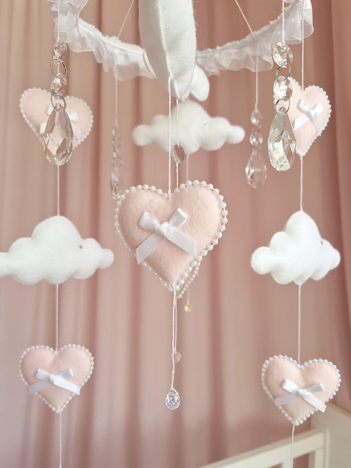 A hanging mobile featuring soft pale pink felt hearts and white felt clouds suspended from a white ruffled circular frame. Each heart is accented with a white satin ribbon bow and a pearl-like beaded trim. Clear crystal-like pendants hang.