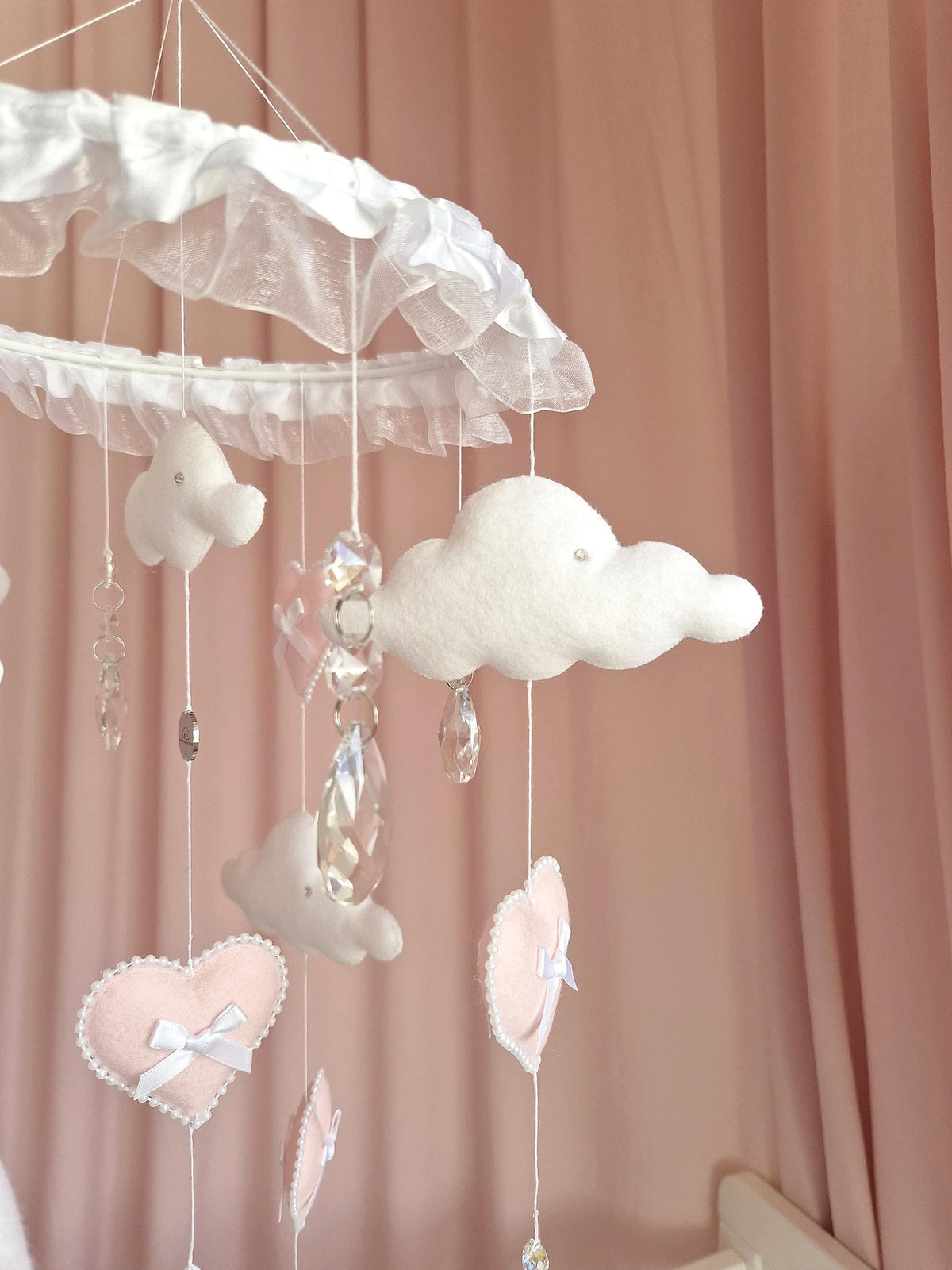 A hanging mobile featuring a white ruffled circular frame with dangling white felt clouds and soft pink felt hearts. The hearts are accented with small white bows and white beaded edges. Clear faceted crystal pendants hang between the shapes.