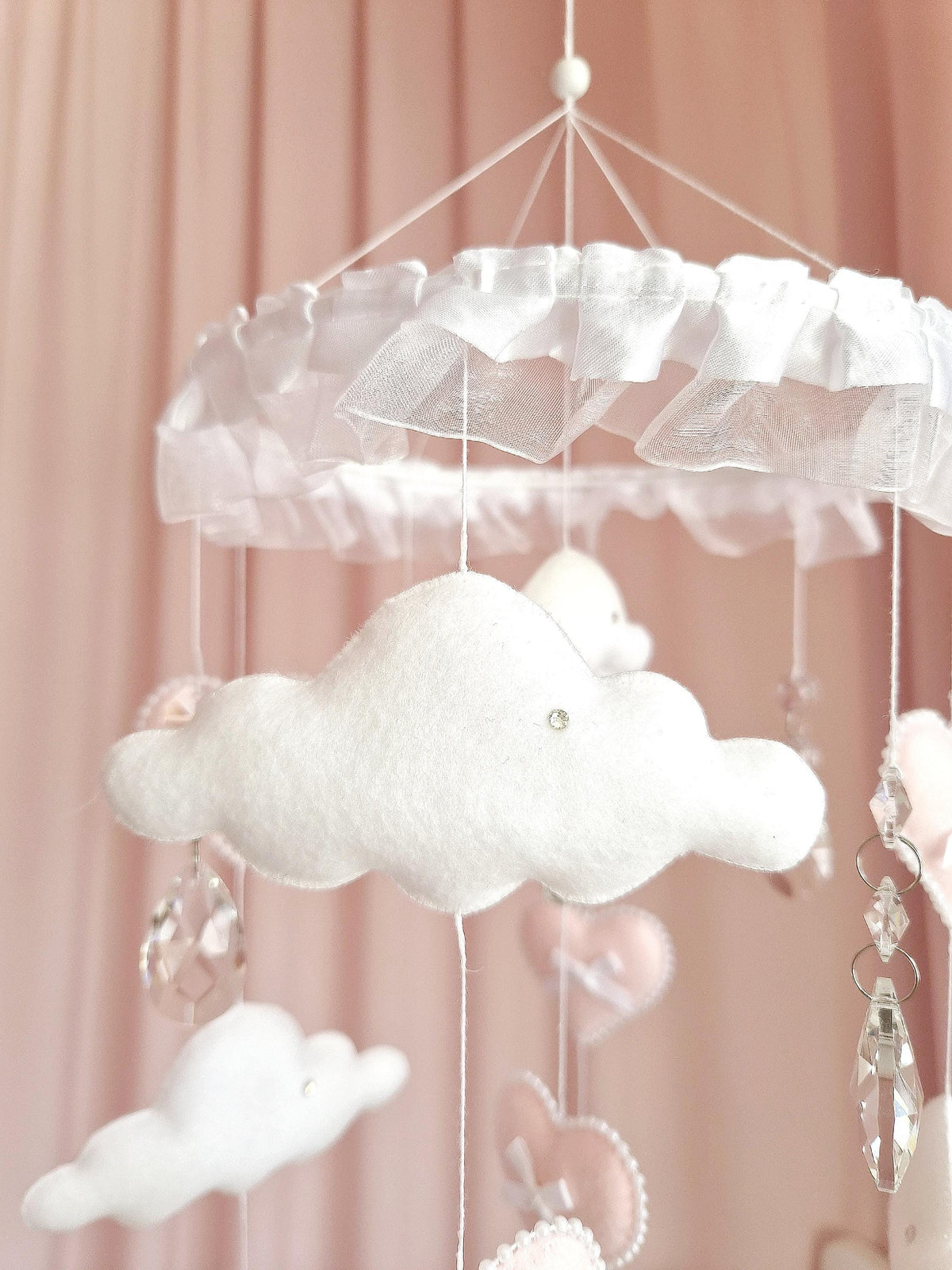 A hanging mobile featuring white felt clouds and soft pink heart-shaped ornaments suspended from a circular frame adorned with white ruffled fabric. Clear crystal-like teardrop pendants hang between the clouds and hearts on thin white strings.