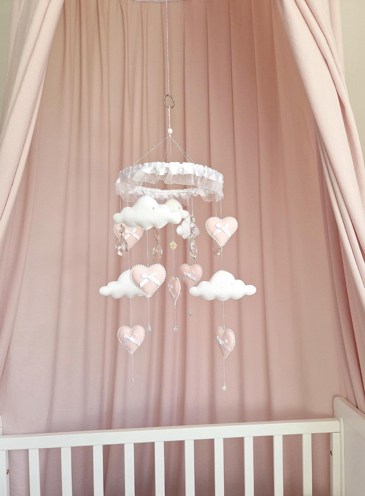 A hanging mobile featuring white fabric clouds and soft pink fabric hearts suspended from a white ruffled circular frame. The mobile includes small white ribbon bows on the hearts and clear crystal-like accents hanging from strings.