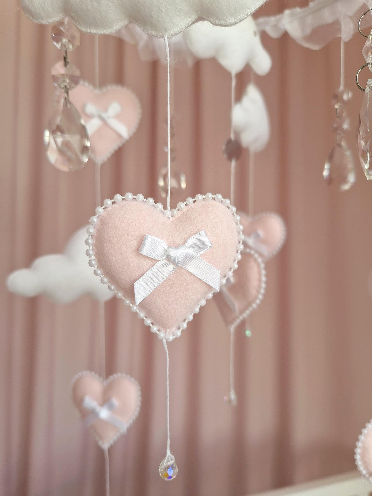 A hanging mobile featuring soft, pale pink felt hearts adorned with white satin bows and delicate pearl-like bead trim. Interspersed among the hearts are white felt clouds and clear, faceted crystal teardrop pendants suspended by white strings.