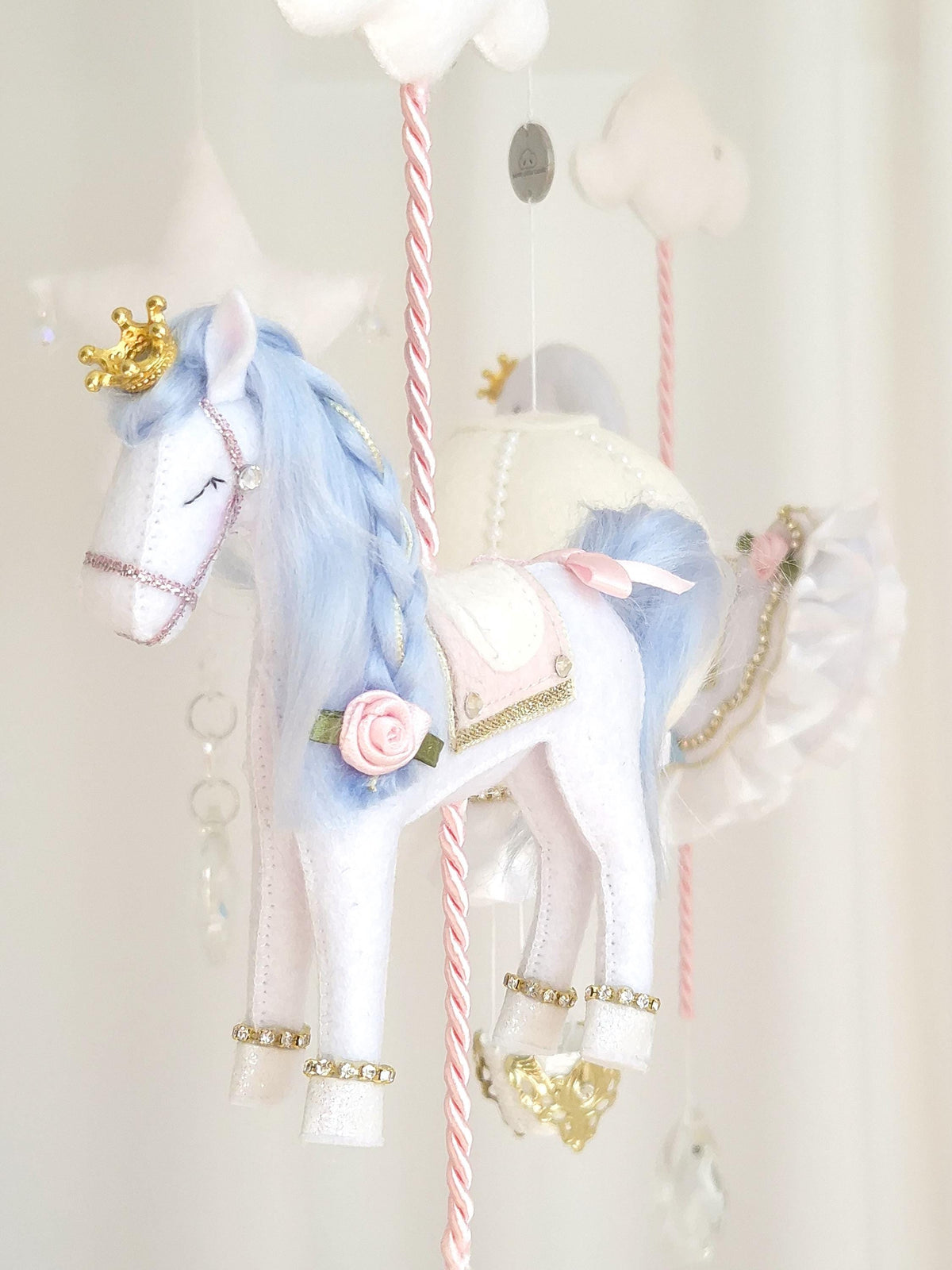 Close-up of a handmade felt carousel horse with blue mane, blush pink flower, gold trim and embroidered details.