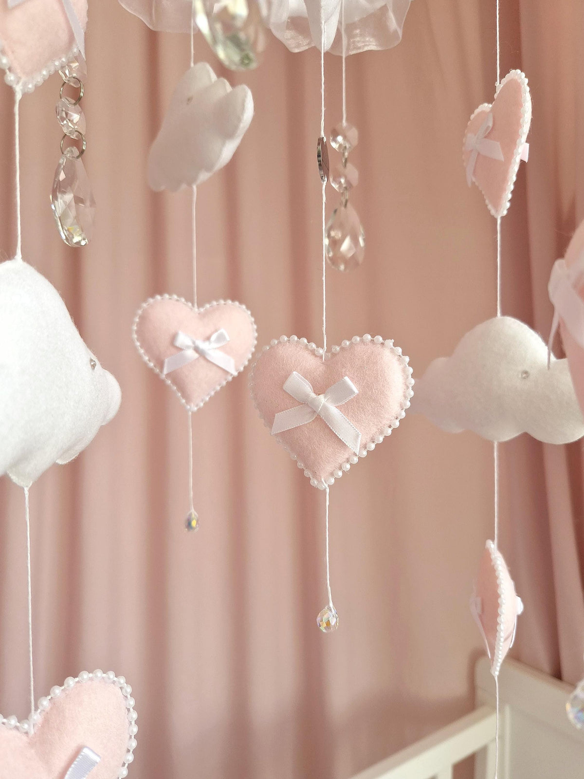 A hanging nursery mobile featuring soft pink felt hearts adorned with white ribbon bows and pearl-like trim, interspersed with white felt clouds and clear faceted crystal pendants suspended on thin white strings against a pink backdrop.
