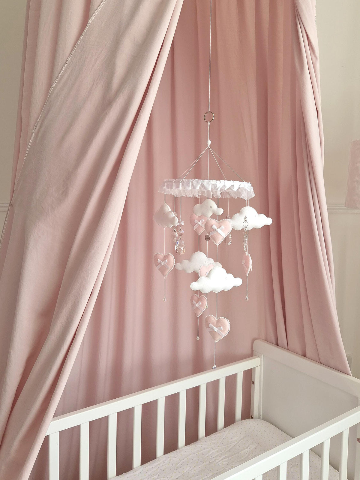 A hanging nursery mobile featuring white felt clouds and soft pink felt hearts suspended from a circular frame with white ruffled trim. The mobile includes small white bows on the hearts and clear crystal-like accents hanging down.