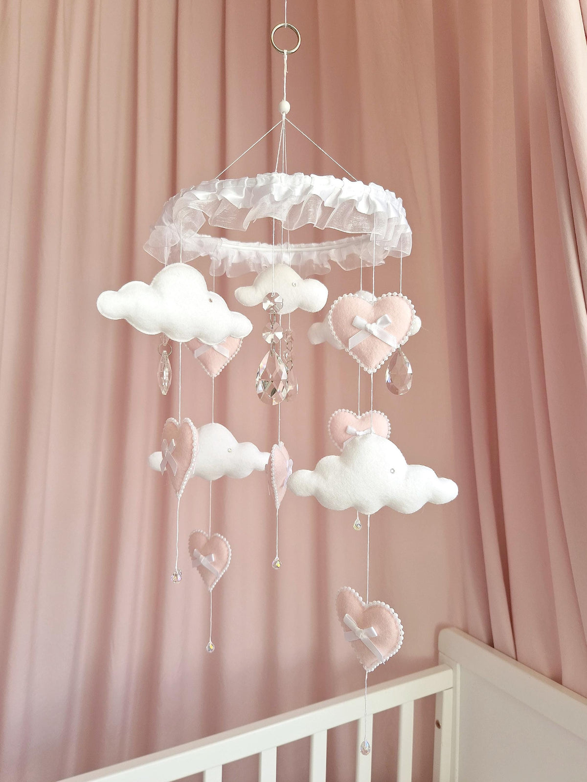 A hanging nursery mobile featuring a white ruffled circular frame with dangling felt clouds and pale pink hearts. The mobile includes white ribbon bows on the hearts and clear faceted crystal teardrop accents suspended by thin white strings.