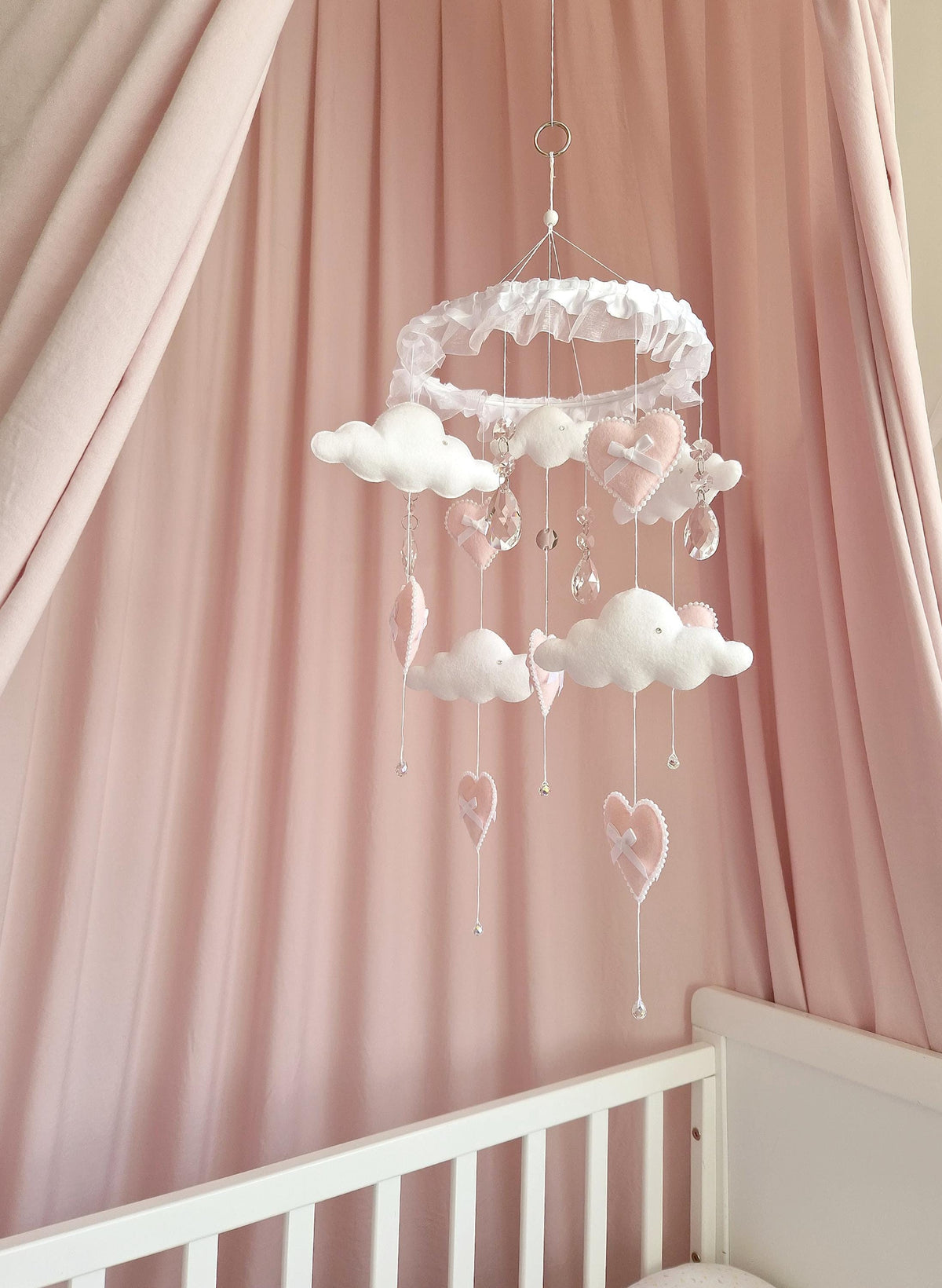 A hanging mobile featuring a white ruffled circular frame with dangling white felt clouds and light pink felt hearts. The mobile includes clear crystal-like teardrop accents and small white beads suspended on thin strings from the frame.
