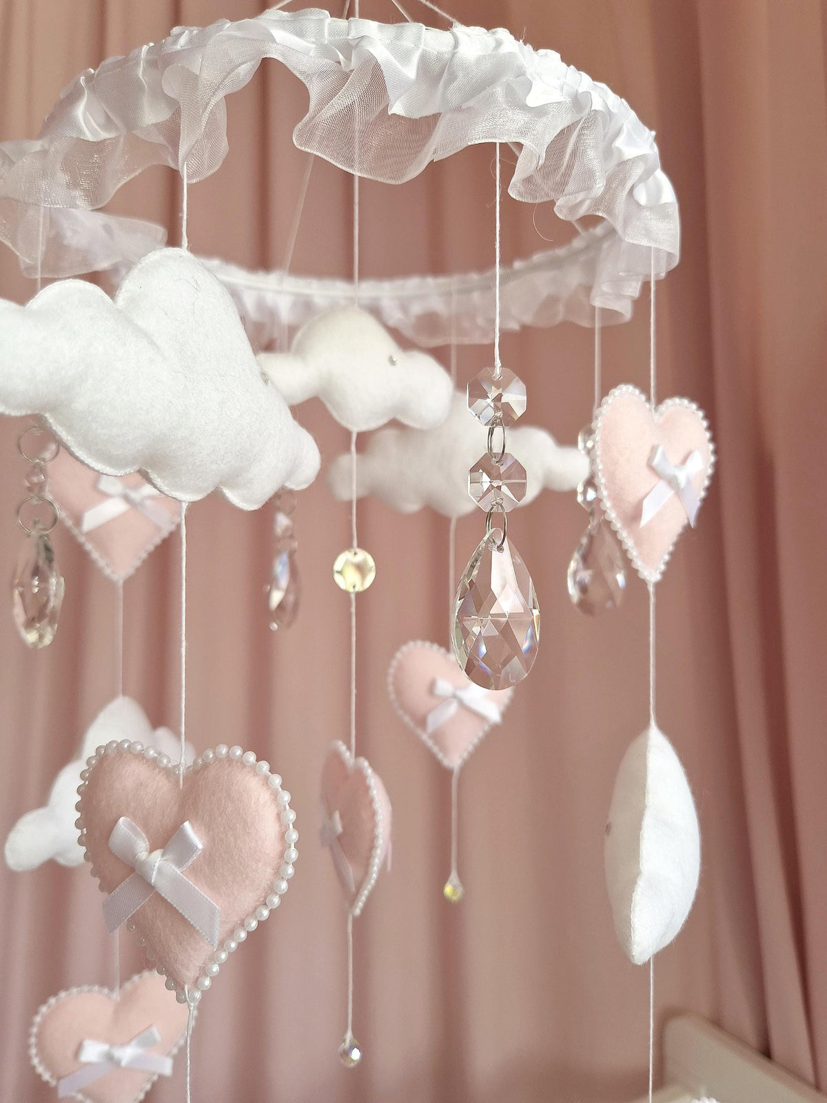 A hanging mobile featuring a white ruffled circular frame with dangling white felt clouds, pale pink felt hearts trimmed with white beads, small white satin bows, and clear faceted crystal pendants suspended on thin white strings.
