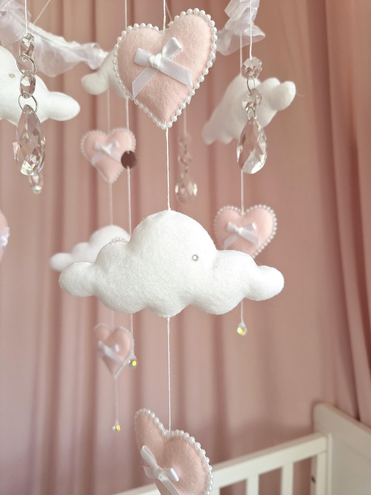 A hanging mobile featuring soft felt hearts in pale pink and white clouds. Each heart is adorned with a small white ribbon bow and a border of tiny white beads. Clear, faceted crystal teardrop pendants hang between the felt shapes.