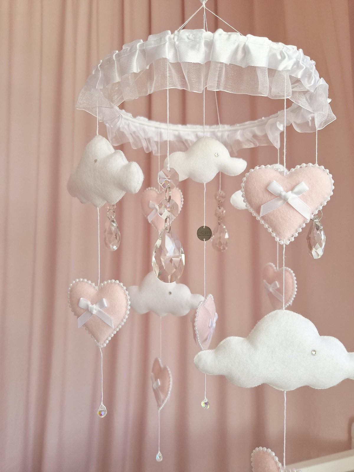 A hanging mobile featuring a white circular frame draped in ruffled fabric. Suspended from the frame are white felt clouds and soft pink felt hearts adorned with small white bows and pearl-like trim, interspersed with clear crystal pendants.