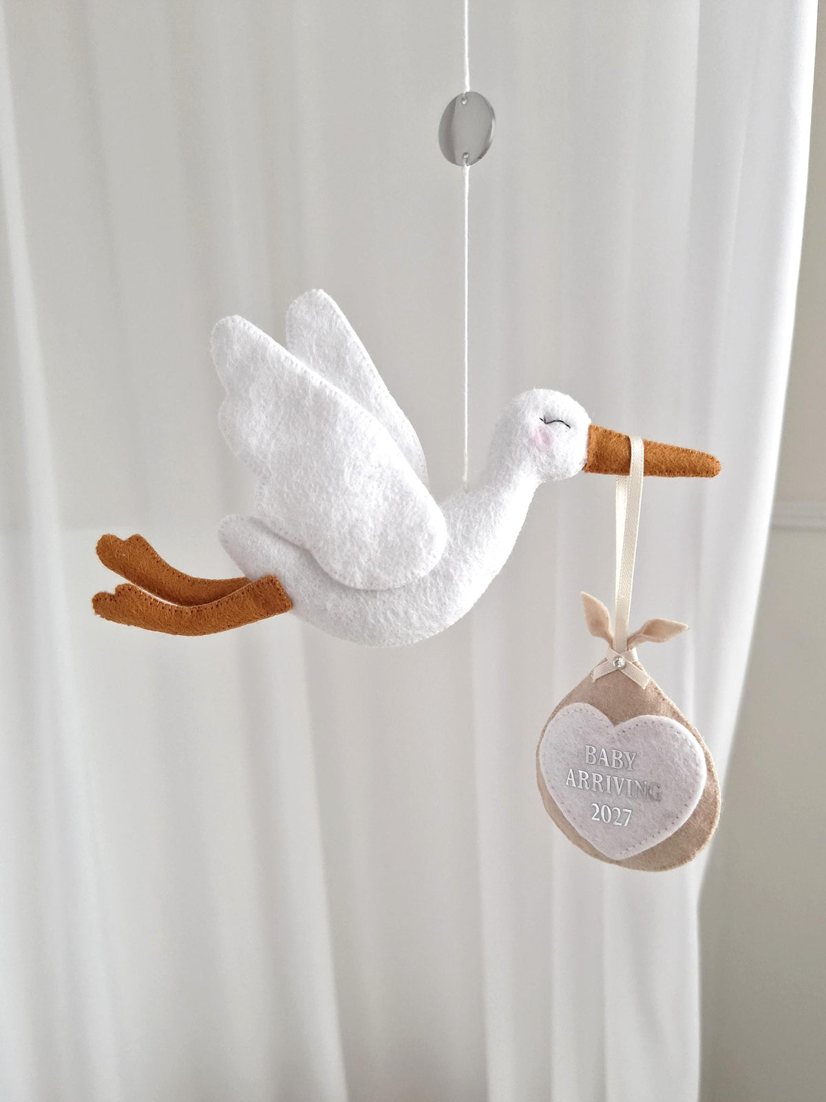 A white felt stork hanging ornament with orange feet and beak, carrying a beige felt bundle suspended by a ribbon. The bundle features a white heart-shaped patch embroidered with the text BABY ARRIVING 2027 in grey thread.