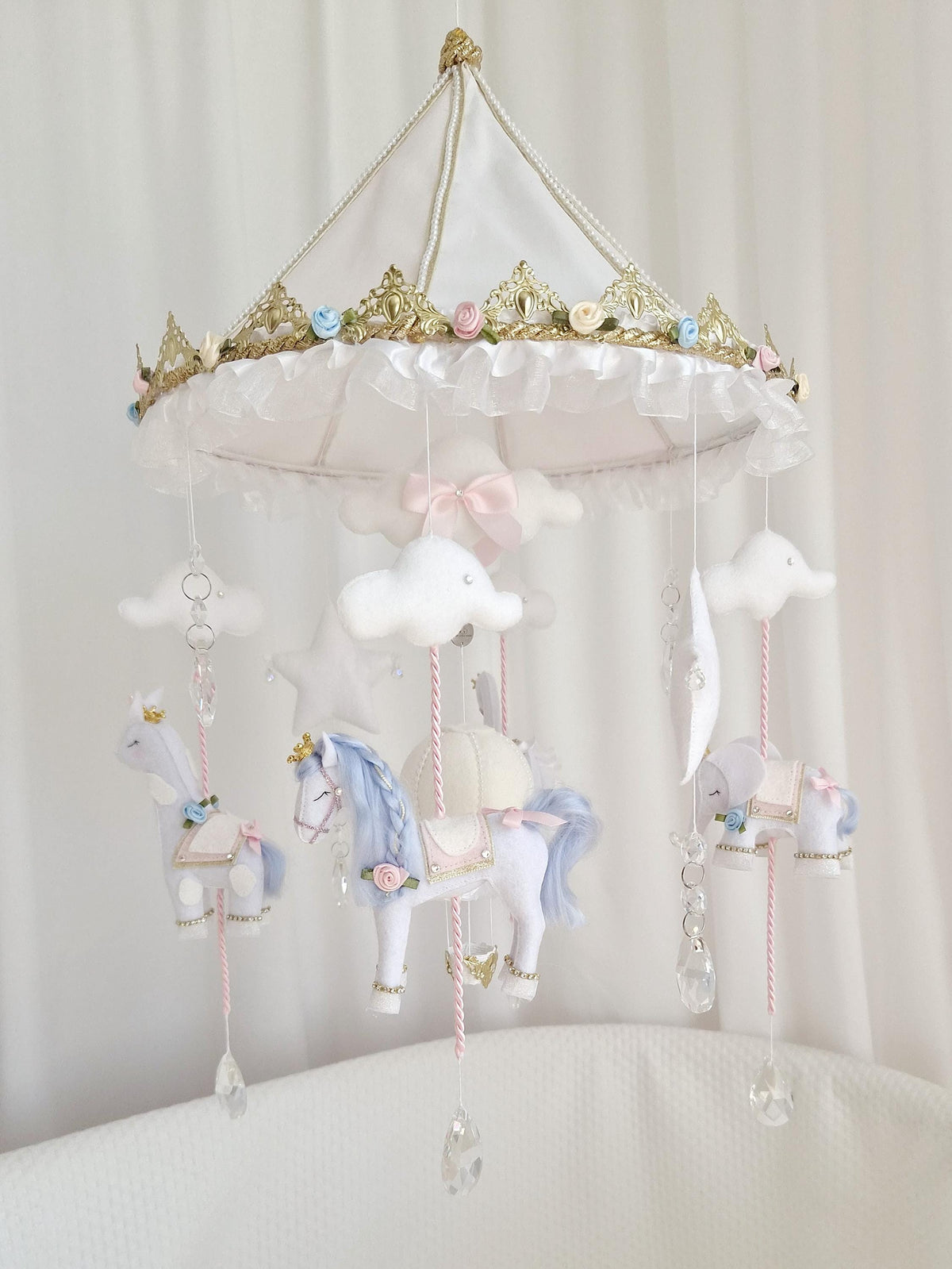 Handmade carousel baby mobile with white, blue and blush pink felt animals, hot air balloon, stars, clouds and glass crystal details, hanging from an ornate gold and white canopy.