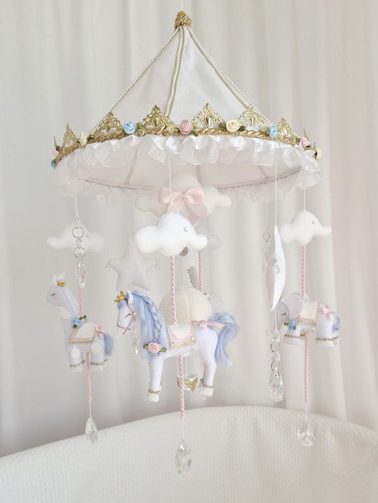 Handmade carousel baby mobile with white, blue and blush pink felt animals, hot air balloon, stars, clouds and glass crystal details, hanging from an ornate gold and white canopy.