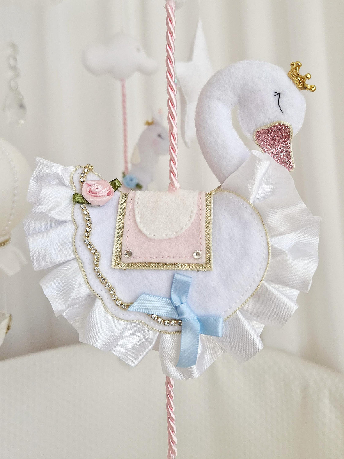 Close-up of a handmade crowned felt swan with layered wings, blush pink saddle and blue ribbon on the carousel nursery mobile.