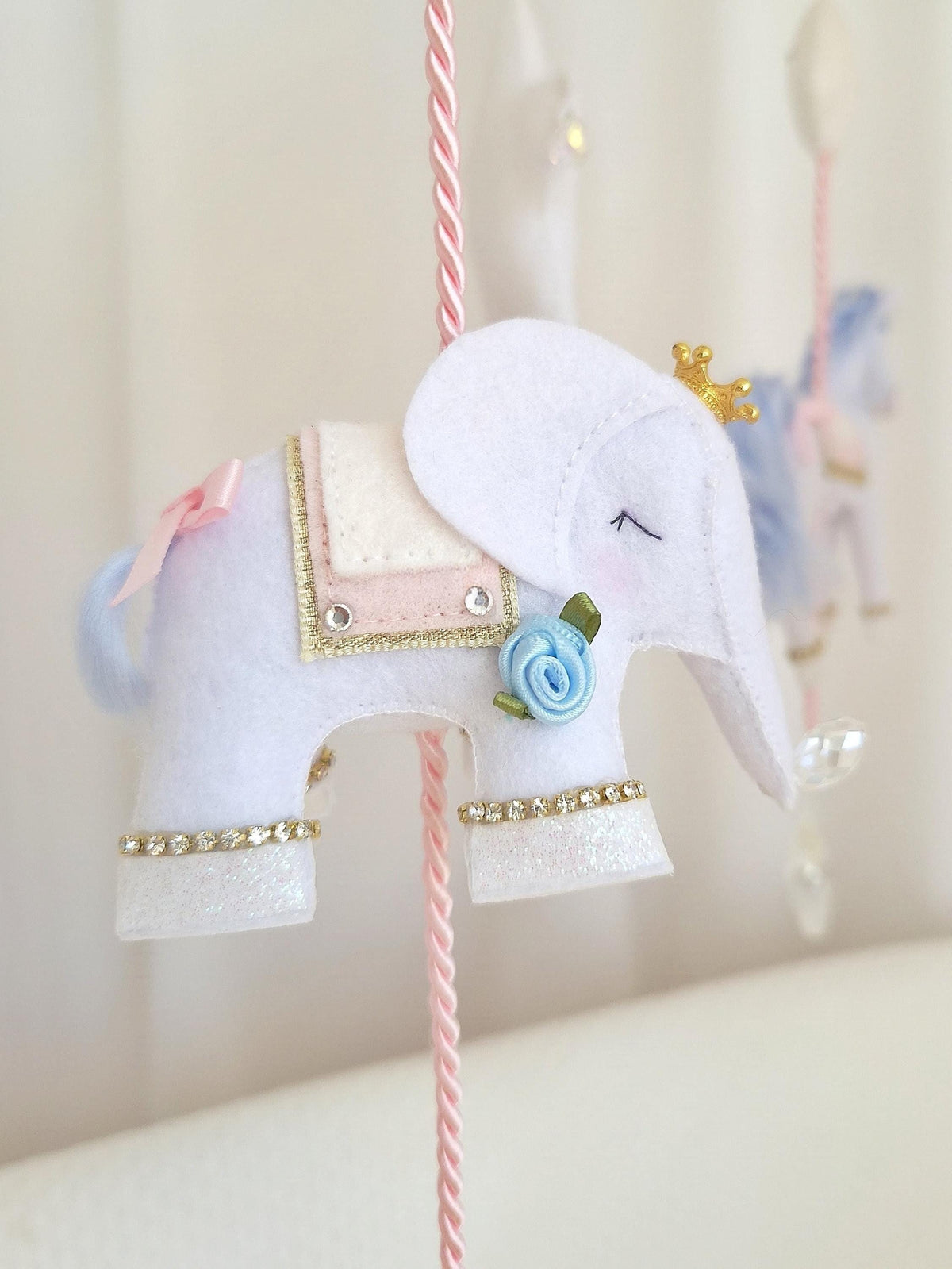 Close-up of a handmade felt carousel elephant with blush pink and blue accents, gold trim and floral embellishments.