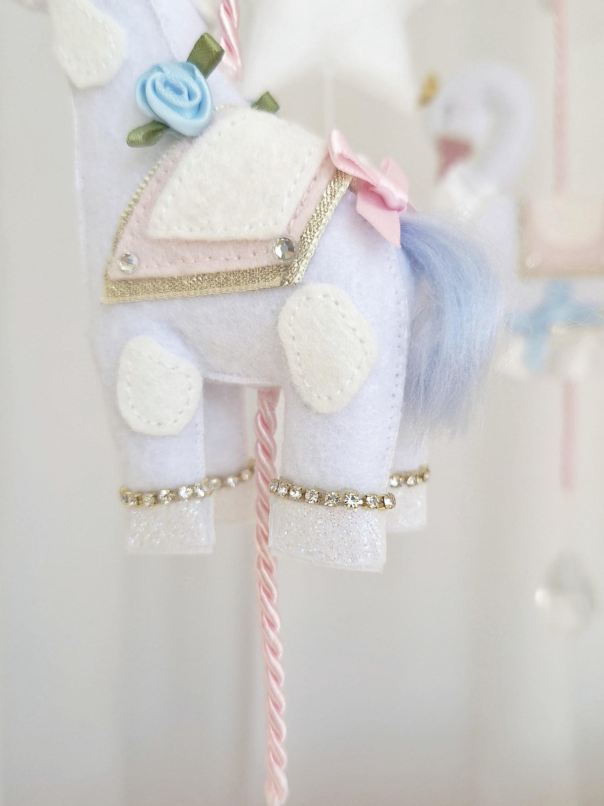Close-up of a handmade felt giraffe with blue mane, blush pink saddle and gold crown hanging from the carousel baby mobile.