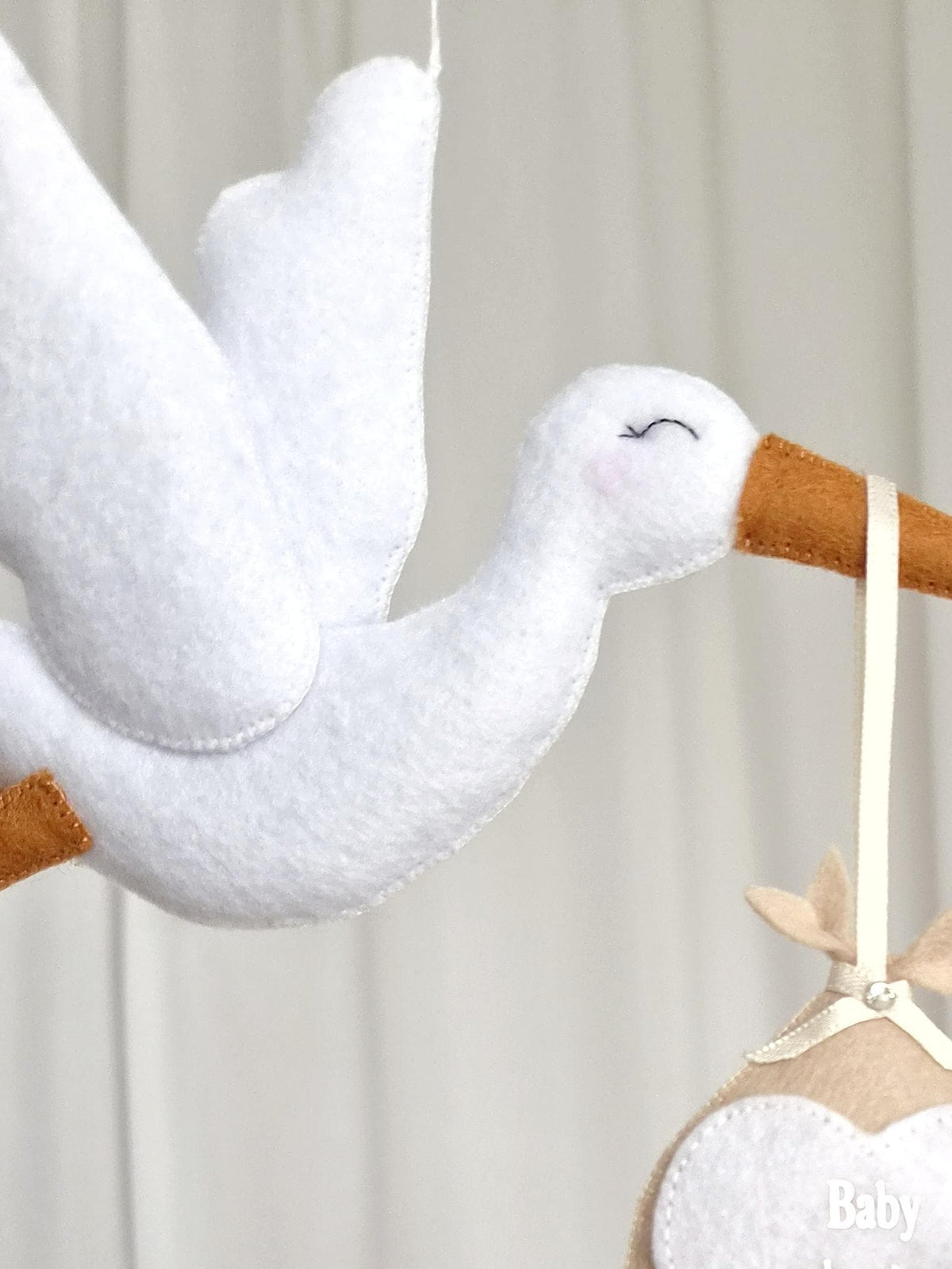 A white felt stork ornament with a tan beak and closed eyes, carrying a tan felt bundle suspended by a cream ribbon. The bundle features a white heart-shaped patch with the word Baby embroidered in white thread on the lower section.