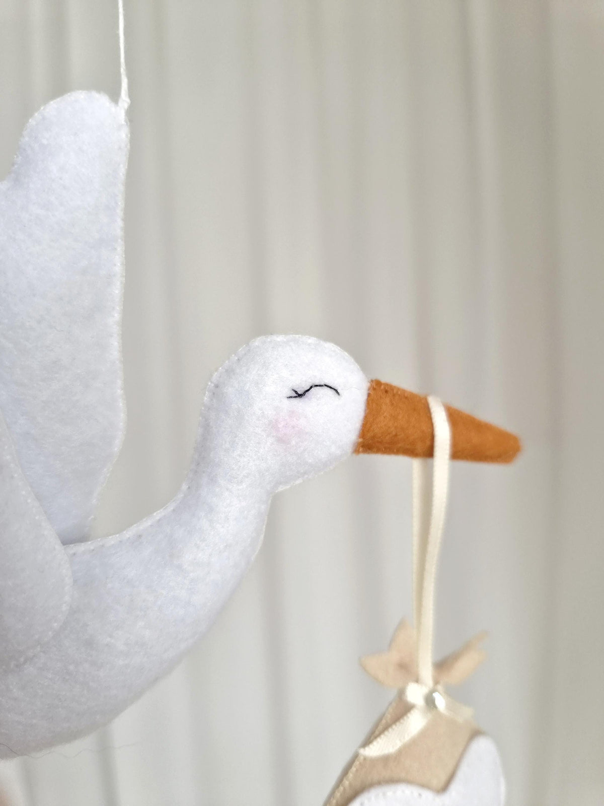 A white felt stork ornament with a brown beak and stitched closed eyes. The stork has a light pink blush on its cheek and holds a cream-coloured ribbon in its beak, which suspends a small, beige, star-shaped felt object below it.