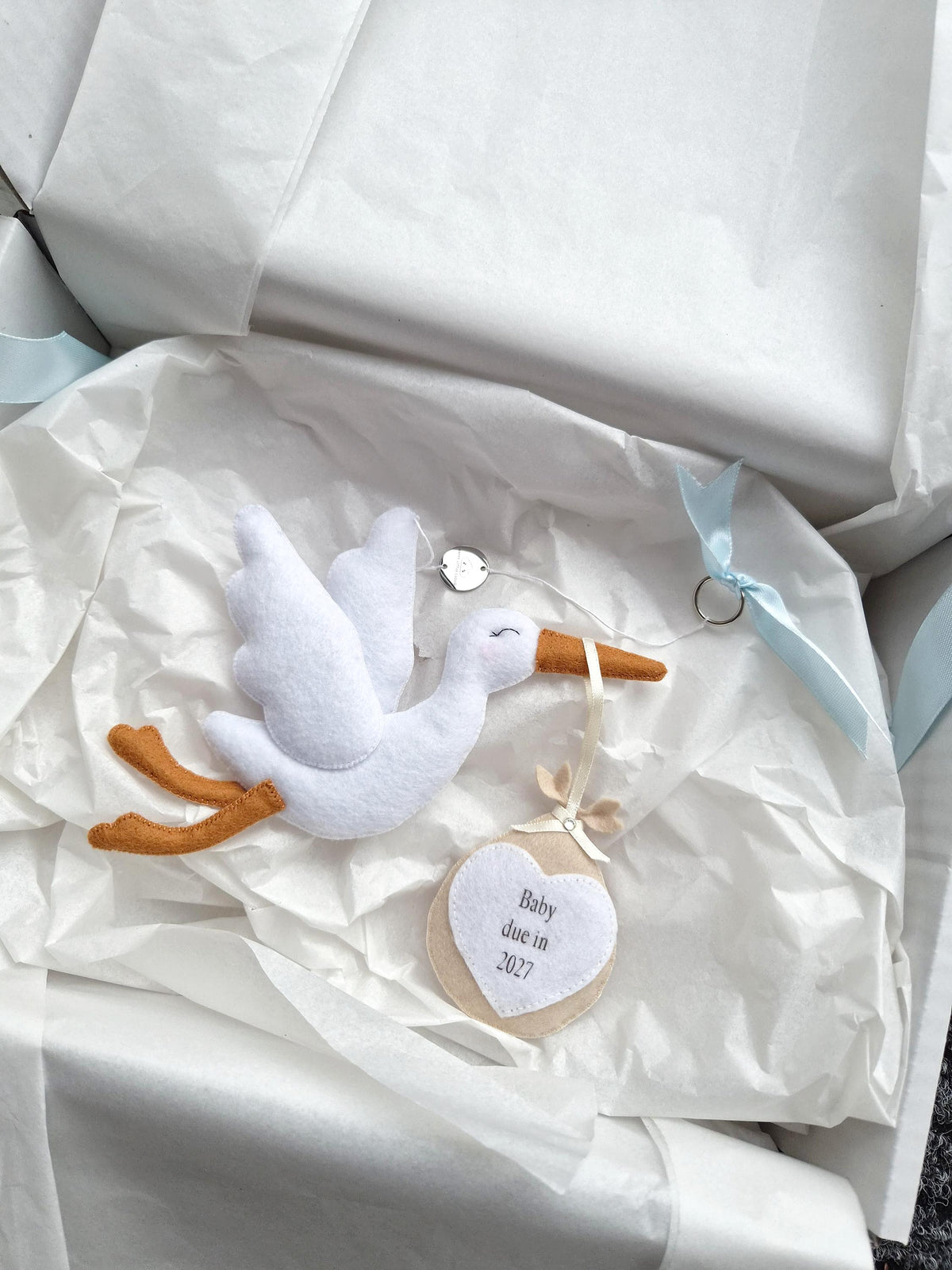 A white felt stork ornament with orange legs and beak, carrying a beige circular bundle with a white heart patch. The heart is embroidered with the text "Baby due in 2027". A light blue ribbon is attached to the top of the ornament.