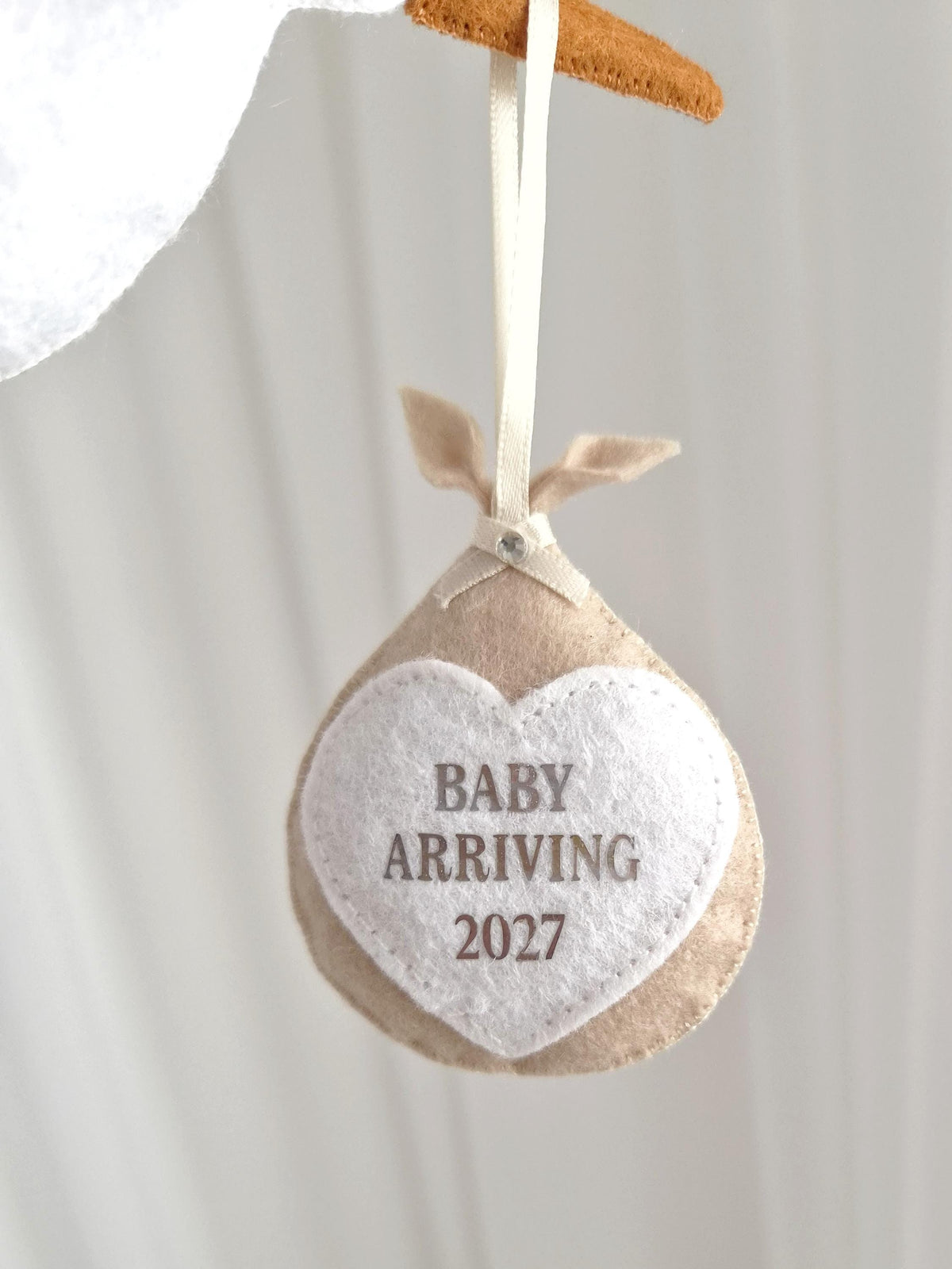 A beige felt bundle ornament hanging from a ribbon, featuring a white heart-shaped patch with the text BABY ARRIVING 2027 in brown lettering. The bundle is tied at the top with a small ribbon and a clear rhinestone accent.