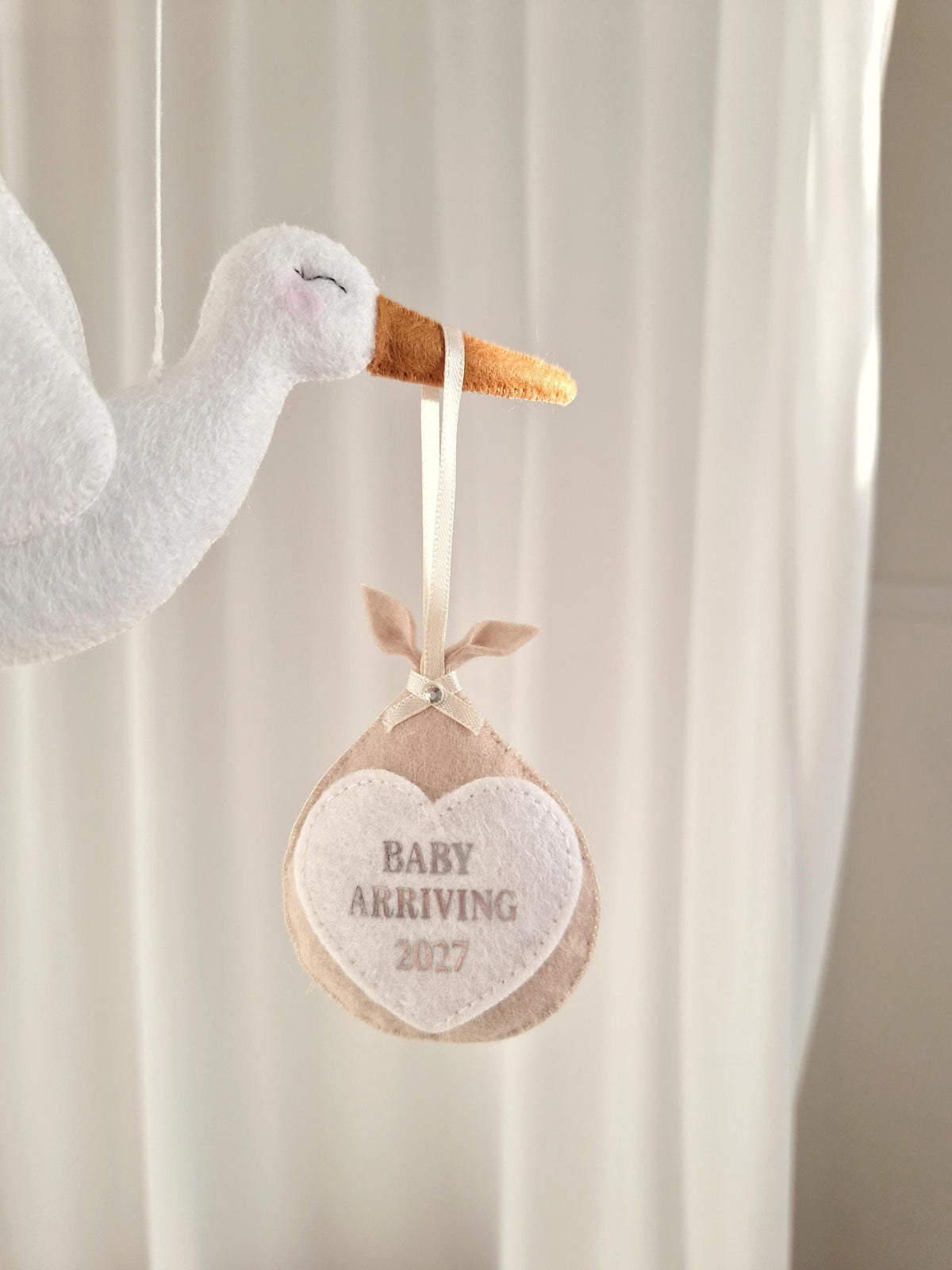 A white felt stork ornament with a brown beak, carrying a beige felt bundle suspended by a cream ribbon. The bundle features a white heart-shaped patch with the embroidered text BABY ARRIVING 2027 in a light grey font.
