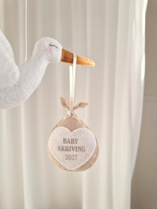 A white felt stork ornament with a brown beak, carrying a beige felt bundle suspended by a cream ribbon. The bundle features a white heart-shaped patch with the embroidered text BABY ARRIVING 2027 in a light grey font.