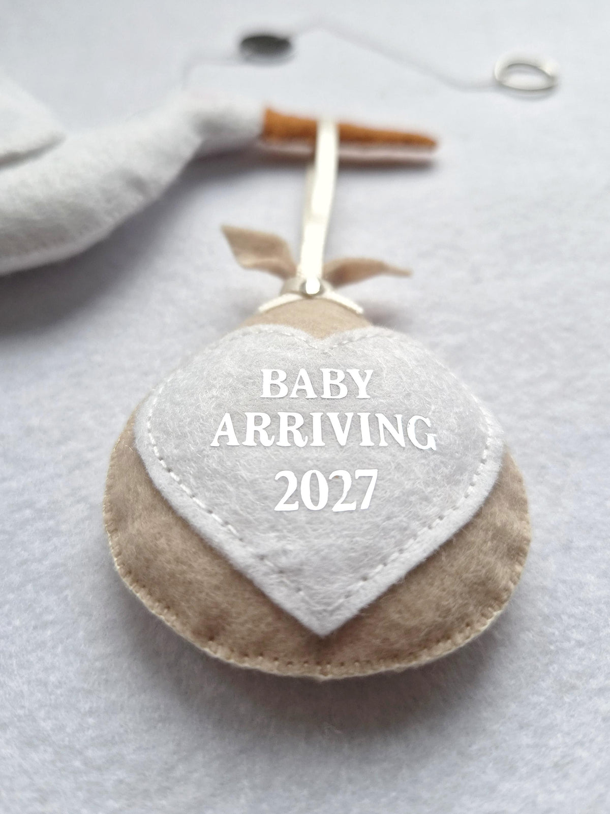 A beige felt bundle ornament featuring a white heart-shaped patch with the text BABY ARRIVING 2027 printed in white. The bundle is suspended by a thin ribbon and is attached to a white felt stork figure with an orange beak.