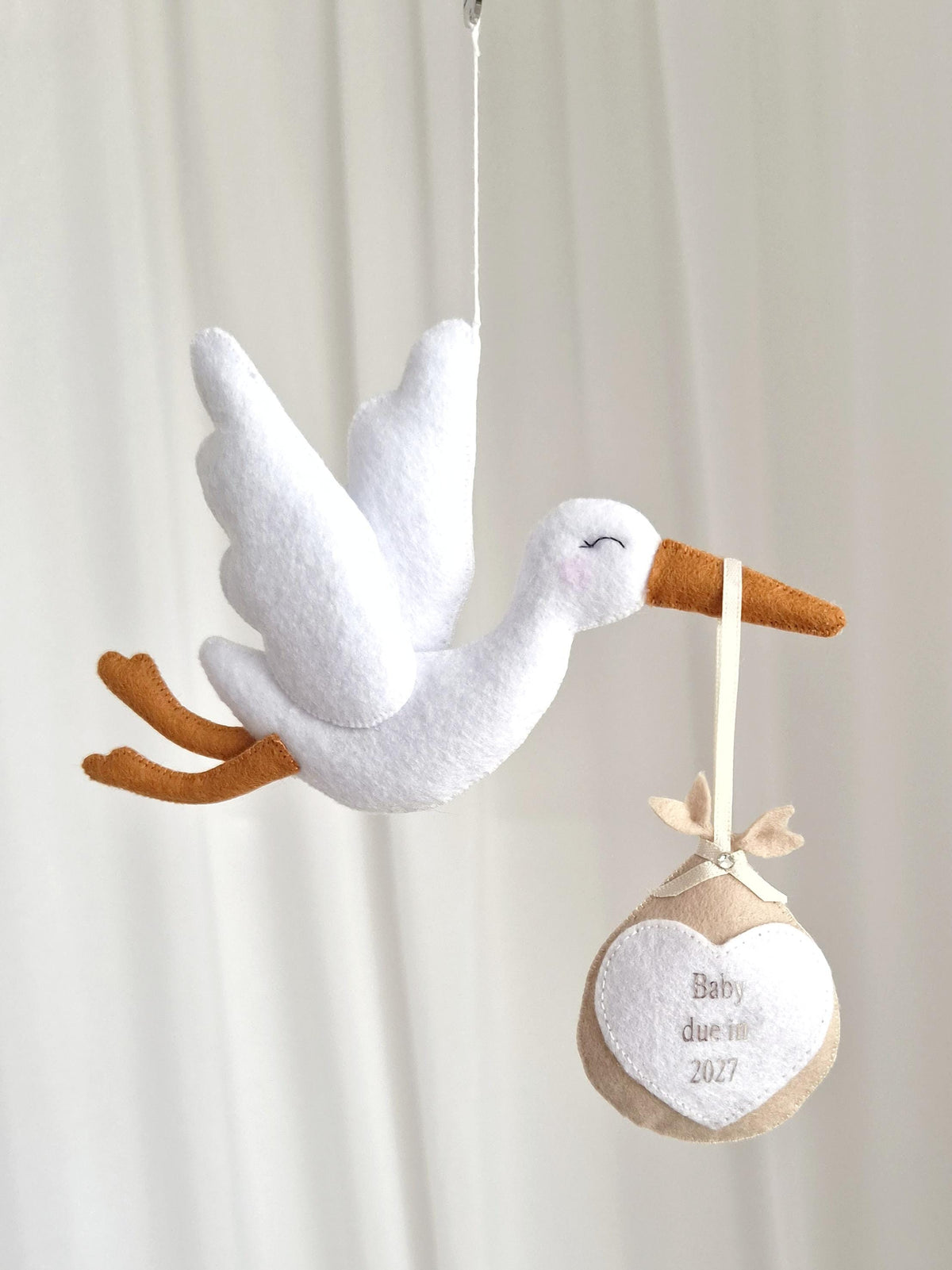 A white felt stork hanging ornament with orange beak and feet, carrying a beige felt bundle suspended by a ribbon. The bundle features a white heart patch with the embroidered text Baby due in 2027, accented by a small clear crystal.