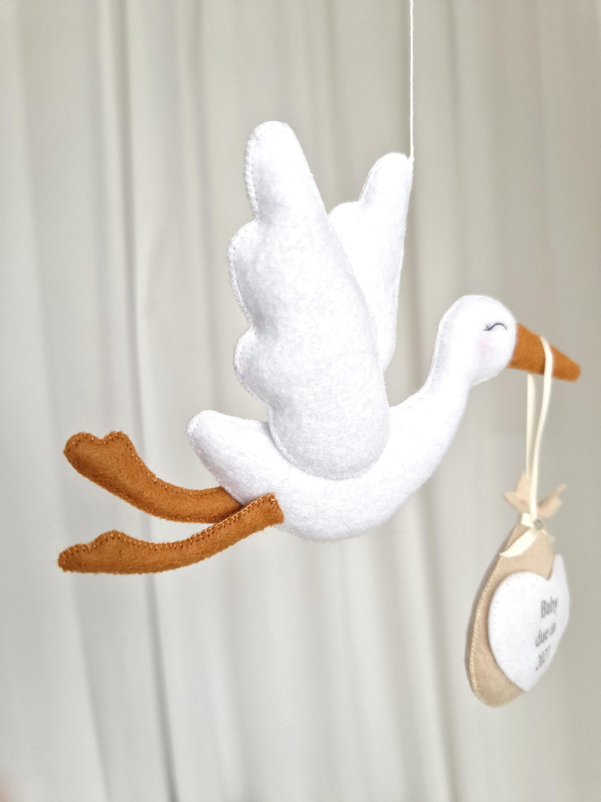 A white felt stork mobile with brown wings and legs, suspended by a thin string. The stork holds a beige fabric bundle with a white heart-shaped tag featuring the text Baby due in 2027, attached by a cream-coloured ribbon.