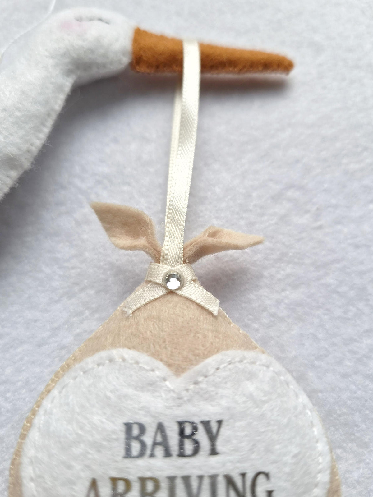 A felt stork ornament with a brown beak, holding a beige felt bundle tied with a cream ribbon and a small clear rhinestone. The bundle features a white heart-shaped patch with the text BABY ARRIVING printed in dark grey letters.