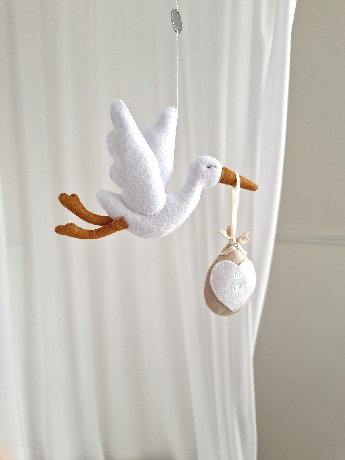A white felt stork mobile with orange legs and beak, suspended by a thin string. The stork carries a beige fabric bundle tied with a ribbon, featuring a white heart-shaped patch with embroidered text on the front of the bundle.
