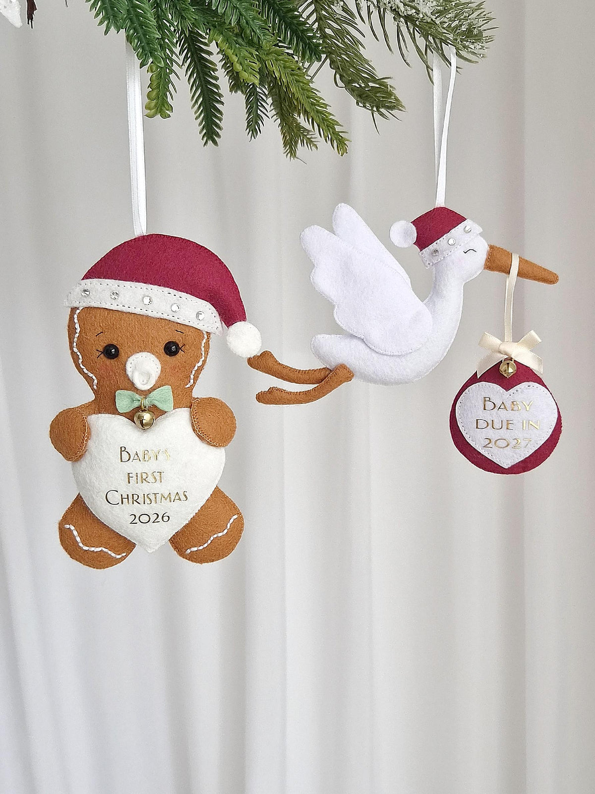 Two felt hanging ornaments: a brown gingerbread person wearing a red Santa hat and a white heart with the text "BABY'S FIRST CHRISTMAS 2026," and a white stork wearing a red Santa hat carrying a red ornament with "BABY DUE IN 2027."