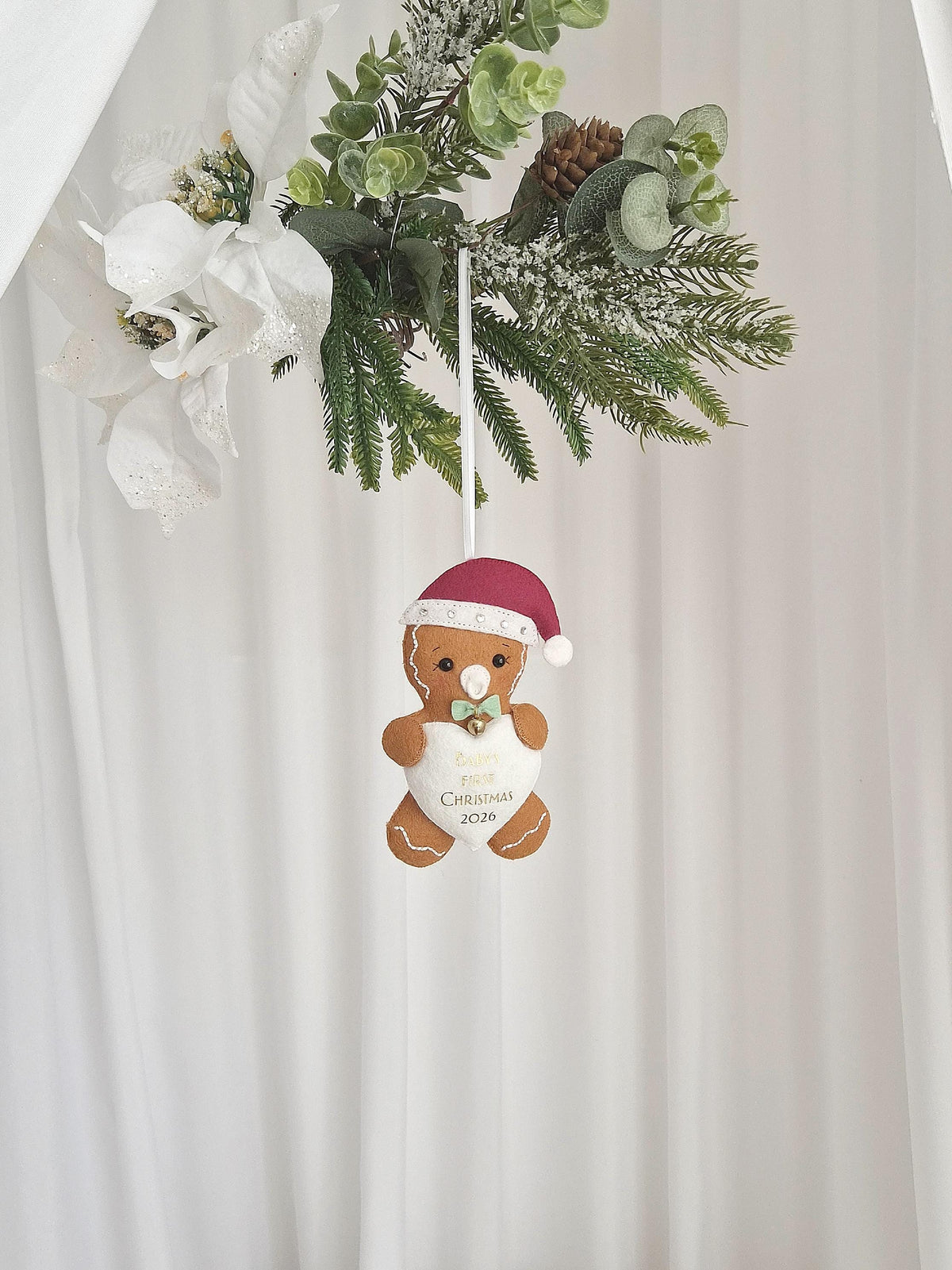 A gingerbread person ornament wearing a maroon Santa hat and a light green bow tie. The figure holds a white heart with the text Baby's First Christmas 2026 printed in gold. The ornament hangs from a white ribbon against a white backdrop.