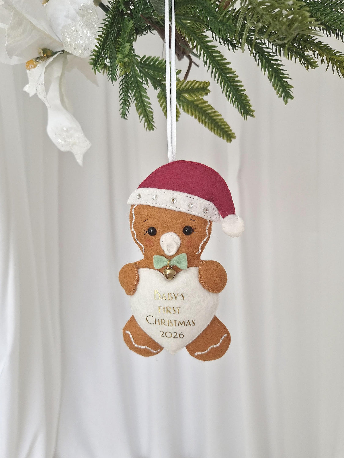 A felt gingerbread person ornament wearing a red Santa hat with rhinestones, a white pacifier, and a light green bow tie with a small bell. It holds a white heart embroidered with the text BABY'S FIRST CHRISTMAS 2026 in gold.