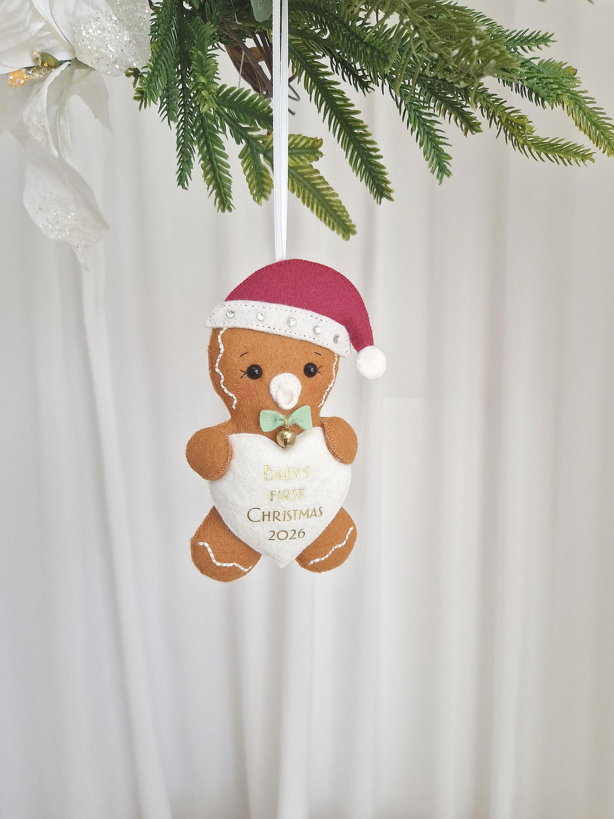 A felt gingerbread person ornament wearing a maroon Santa hat with white trim and a light green bow tie. The figure holds a white heart embroidered with the text Baby's First Christmas 2026 and features a small gold bell.