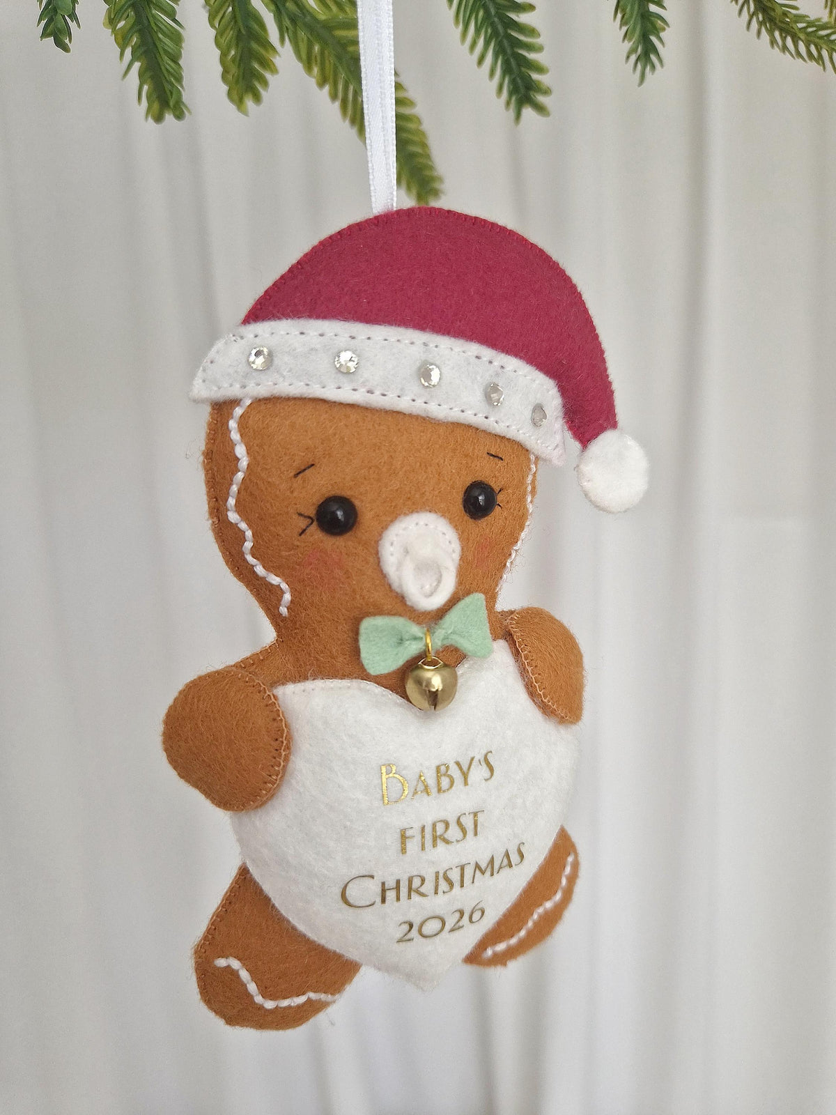 A felt gingerbread person ornament wearing a red Santa hat with rhinestones, a white pacifier, and a light green bow tie with a gold bell. A white heart on the chest features the gold text BABY'S FIRST CHRISTMAS 2026. It hangs by a white ribbon.