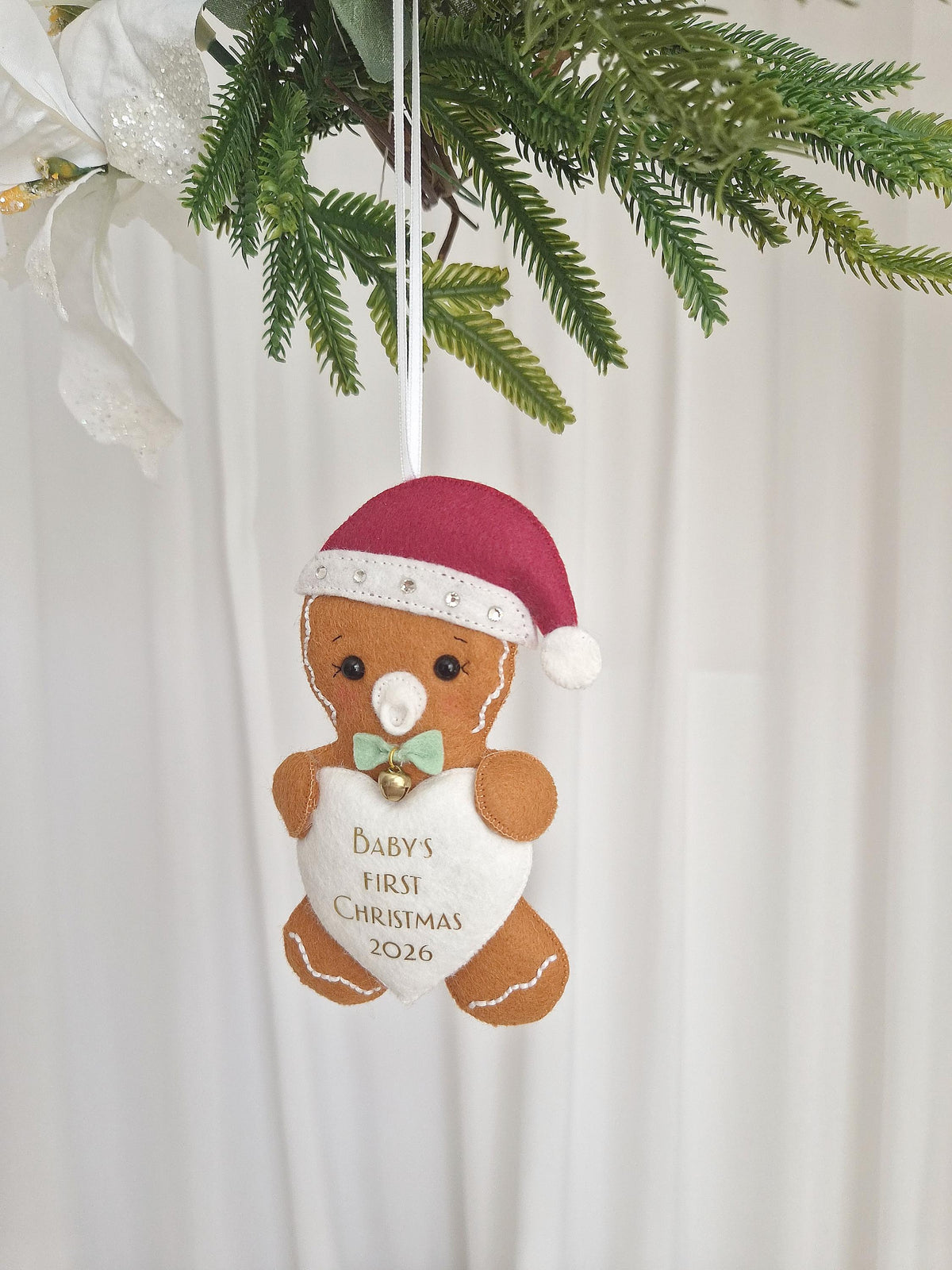 A felt gingerbread person ornament wearing a maroon Santa hat with silver rhinestones, a light green bow tie with a small gold bell, and a white heart-shaped patch on the chest featuring the text Baby's First Christmas 2026.