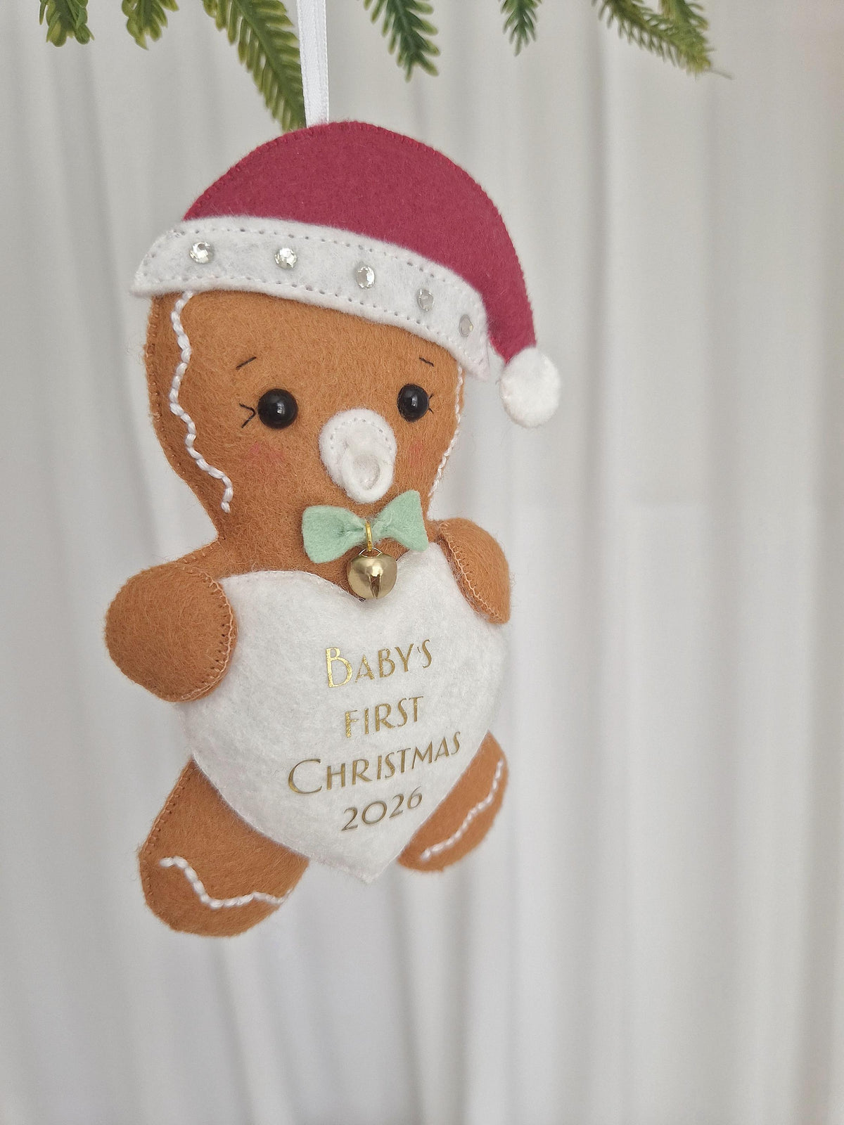 A felt gingerbread person ornament wearing a red Santa hat with white trim and rhinestones. The figure has a white dummy, a light green bow tie with a small gold bell, and a white heart-shaped patch with the gold text BABY'S FIRST CHRISTMAS 2026.