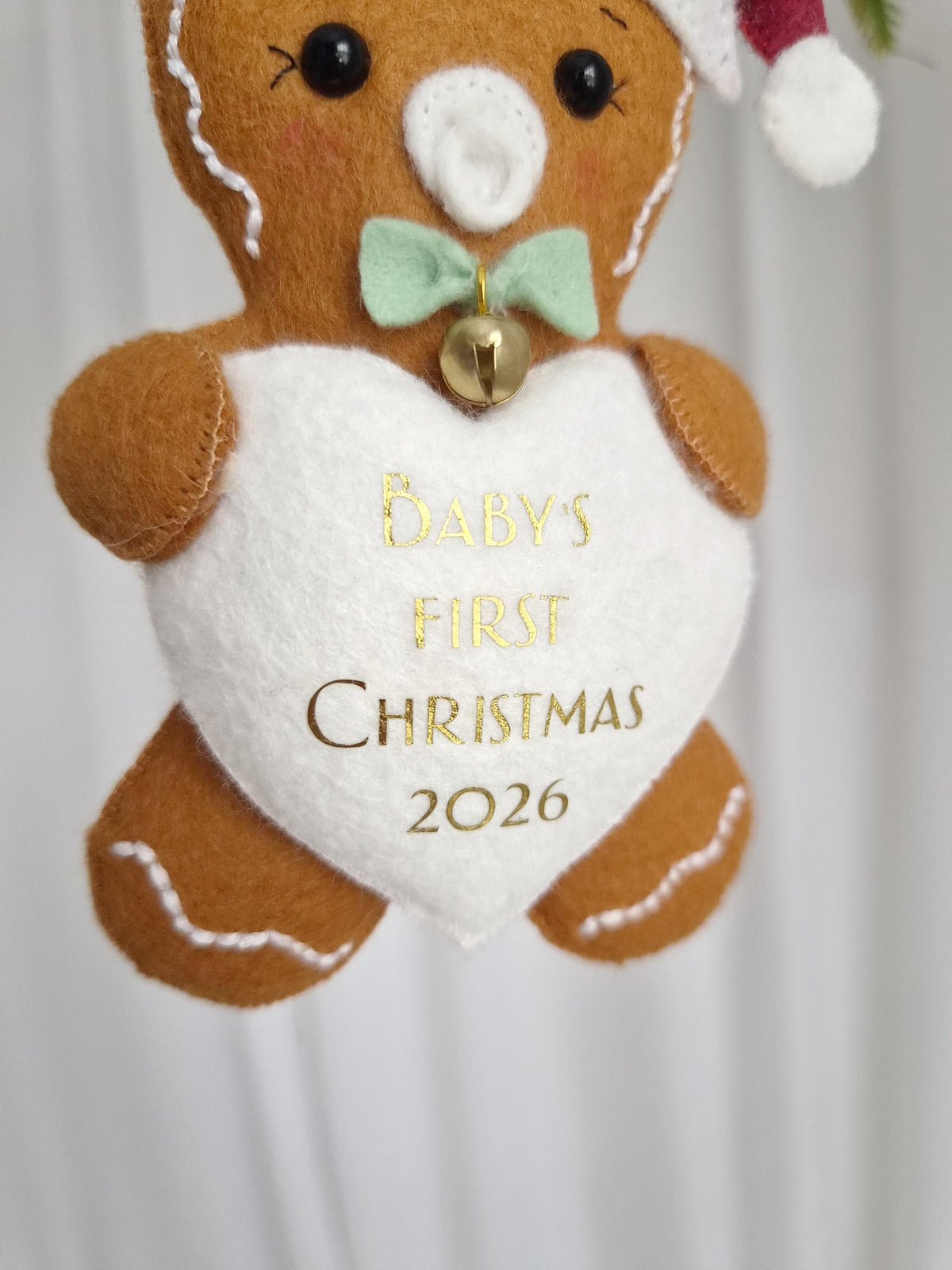 A brown felt gingerbread bear ornament wearing a white and red hat, a light green bow tie with a small gold bell, and holding a white heart-shaped patch. The heart features gold text reading BABY'S FIRST CHRISTMAS 2026.