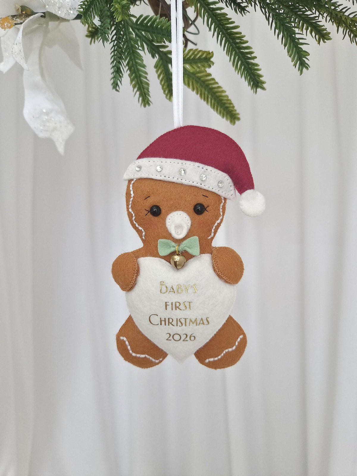 A felt gingerbread person ornament wearing a maroon Santa hat with white trim and rhinestones. It has a white pacifier, a light green bow tie with a small gold bell, and holds a white heart embroidered with "BABY'S FIRST CHRISTMAS 2026".