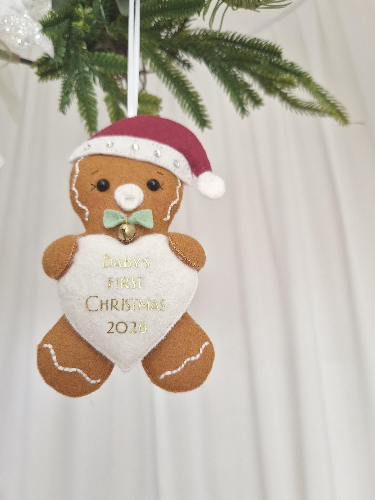 A felt gingerbread person ornament wearing a maroon Santa hat with white trim and a light green bow with a small gold bell. The figure holds a white heart embroidered with the gold text BABY'S FIRST CHRISTMAS 2026.