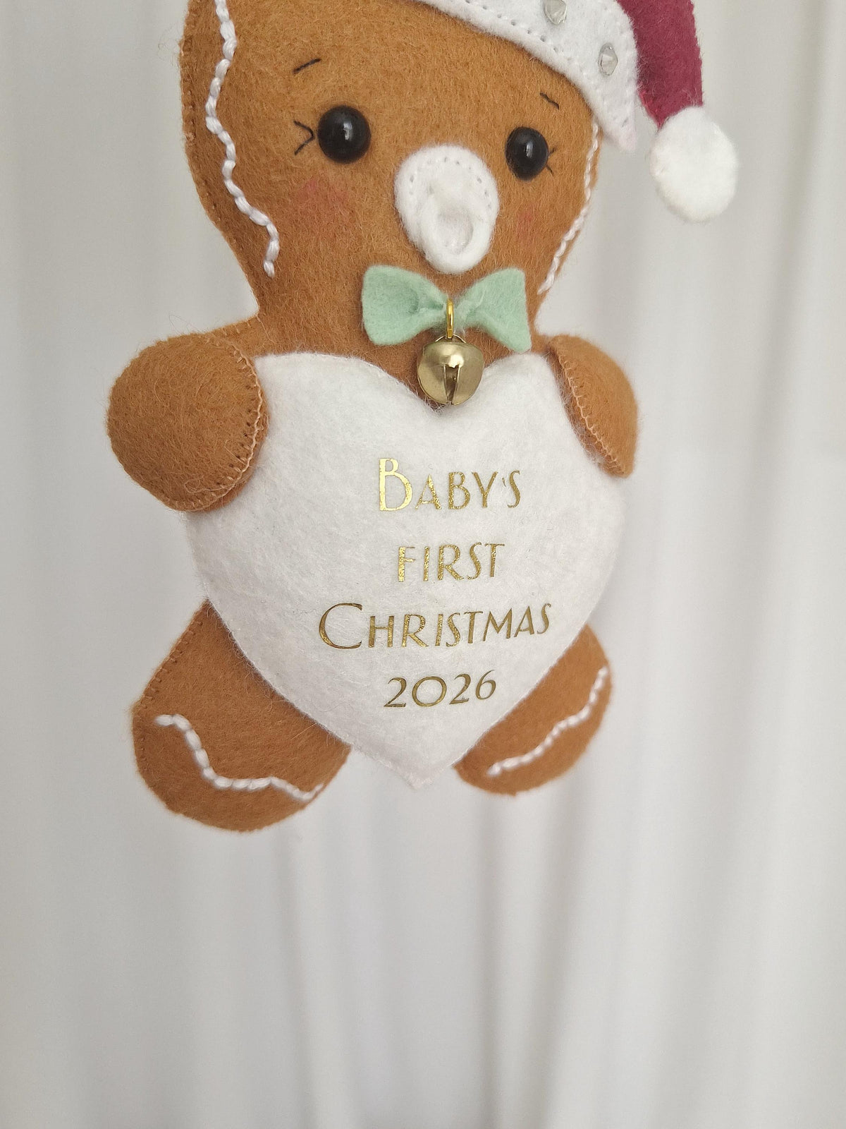 A brown felt gingerbread ornament featuring a white heart with the gold text BABY'S FIRST CHRISTMAS 2026. The figure wears a red and white Santa hat, a light green bow tie with a small gold bell, and has a white pacifier in its mouth.