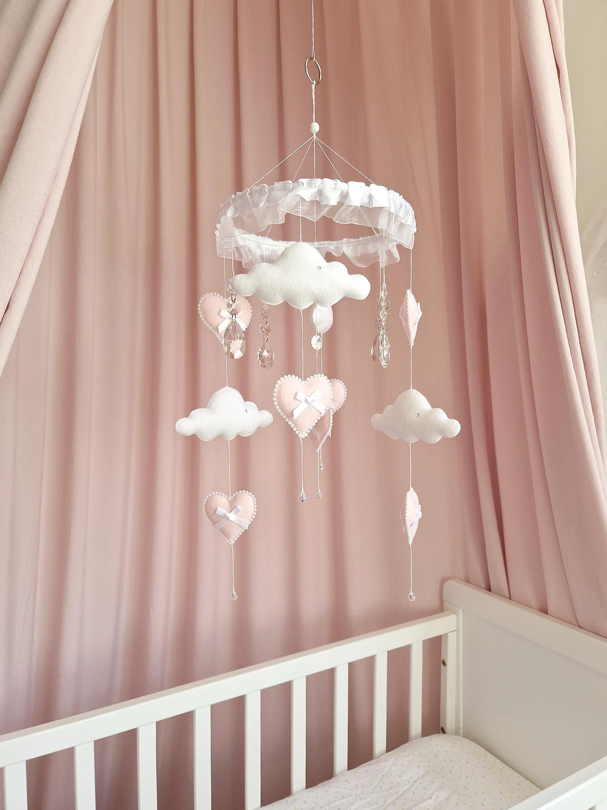 A hanging nursery mobile featuring a white circular frame with ruffled trim, suspending soft white clouds and pale pink heart-shaped ornaments. The mobile includes clear crystal-like teardrop accents and small white bows attached to the hearts.