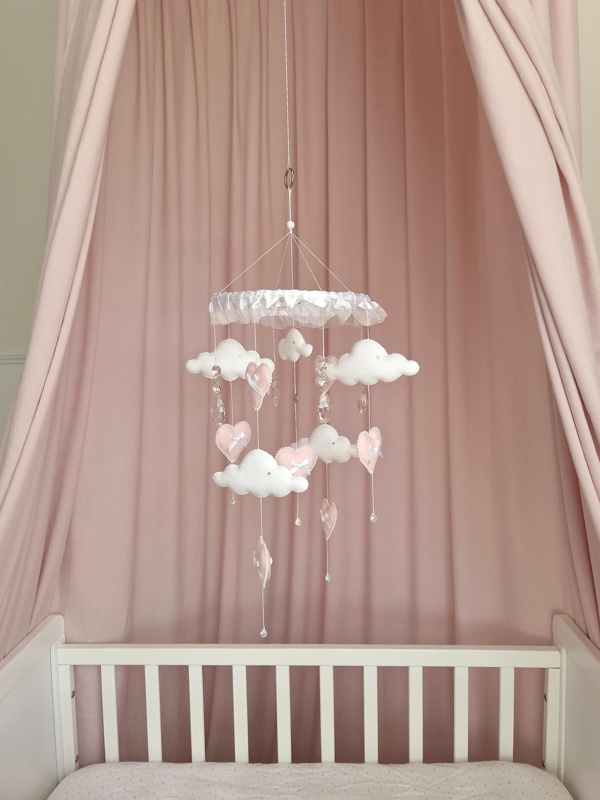 A hanging nursery mobile featuring white fabric clouds and soft pink fabric hearts suspended from a circular frame. The mobile includes clear crystal-like accents hanging on thin white strings, creating a layered, delicate arrangement.