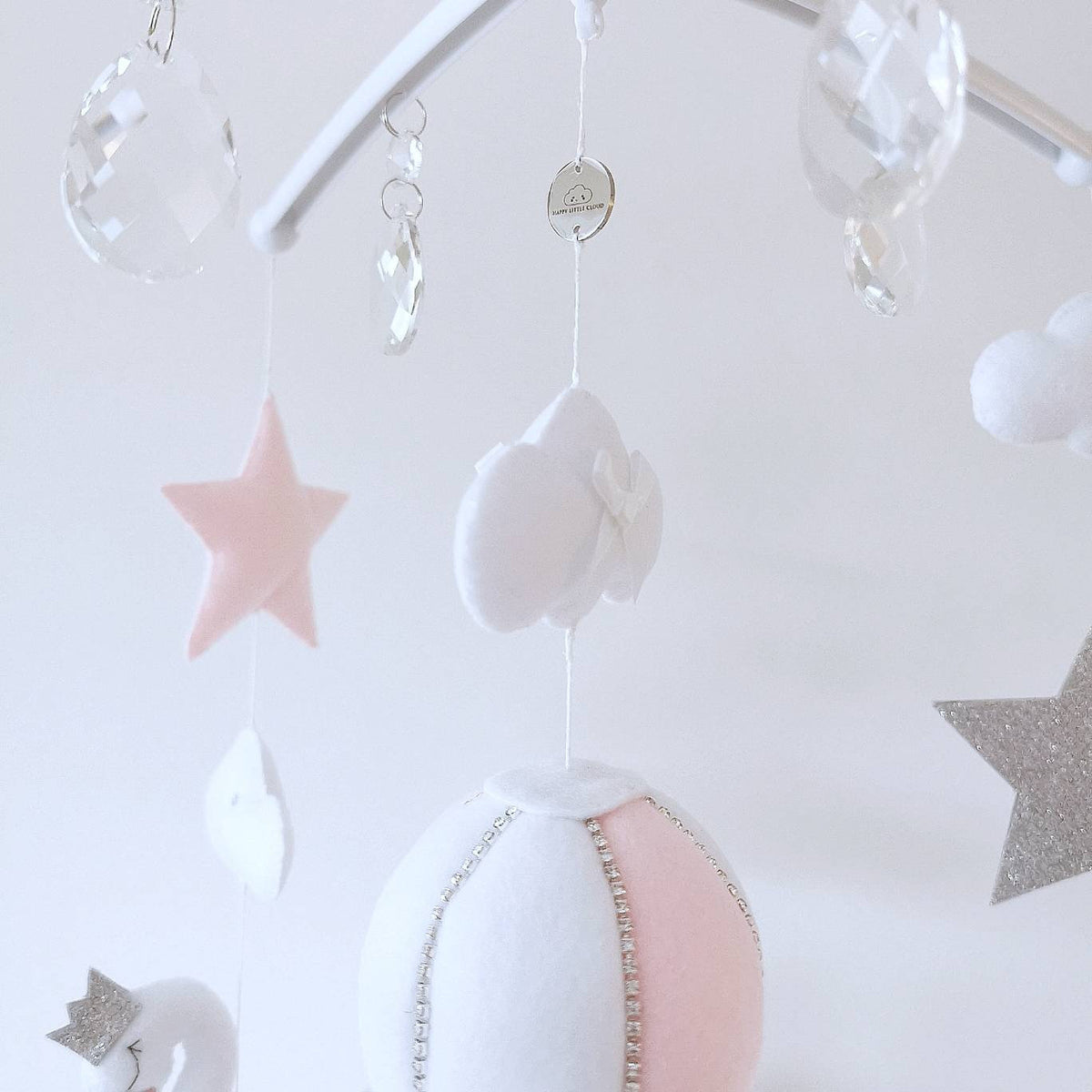 A softly stitched blush balloon floats in the centre of the mobile surrounded by gentle shapes and shimmering details

