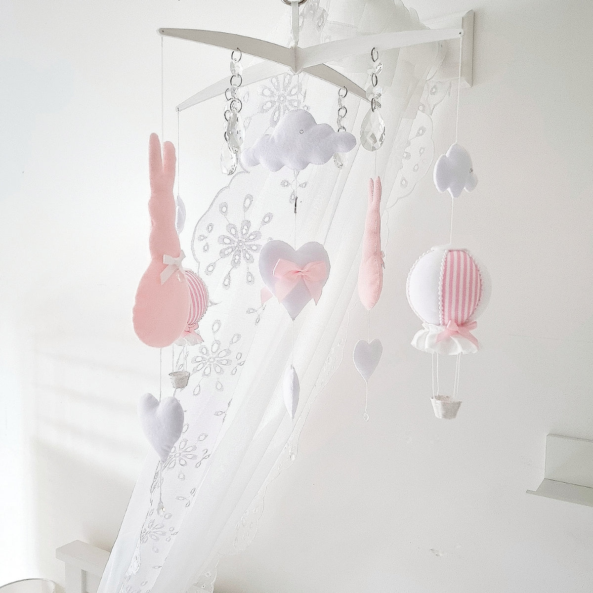 Soft handmade baby girl mobile with pink bunny, heart, cloud, and balloon elements hanging from a wooden cross hanger.